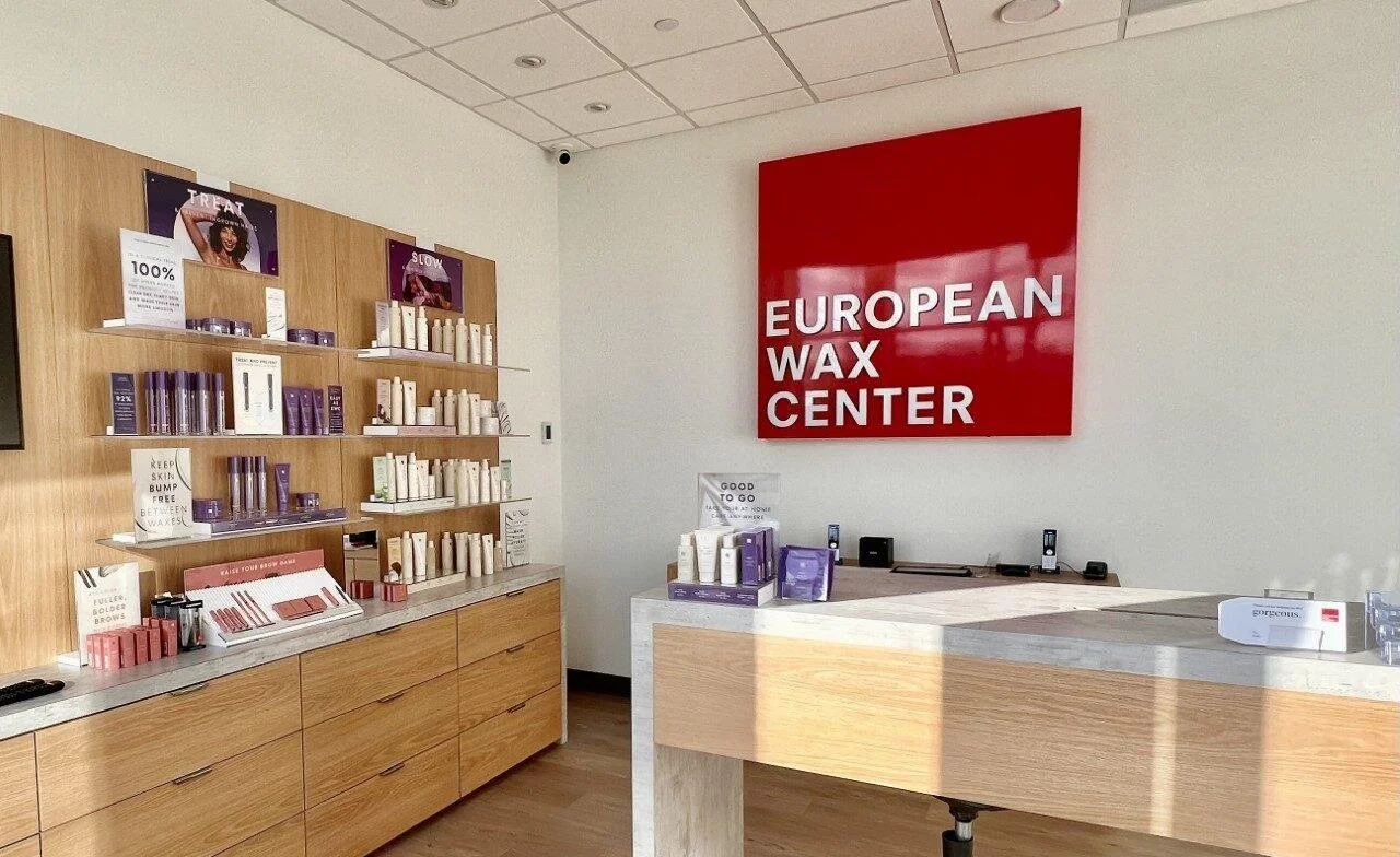 Interior of a European wax center with wooden shelves displaying skincare products. A bold red sign reads 'European Wax Center' on the white wall, and a wooden reception counter is in the foreground. HVAC Services New York City, USA.