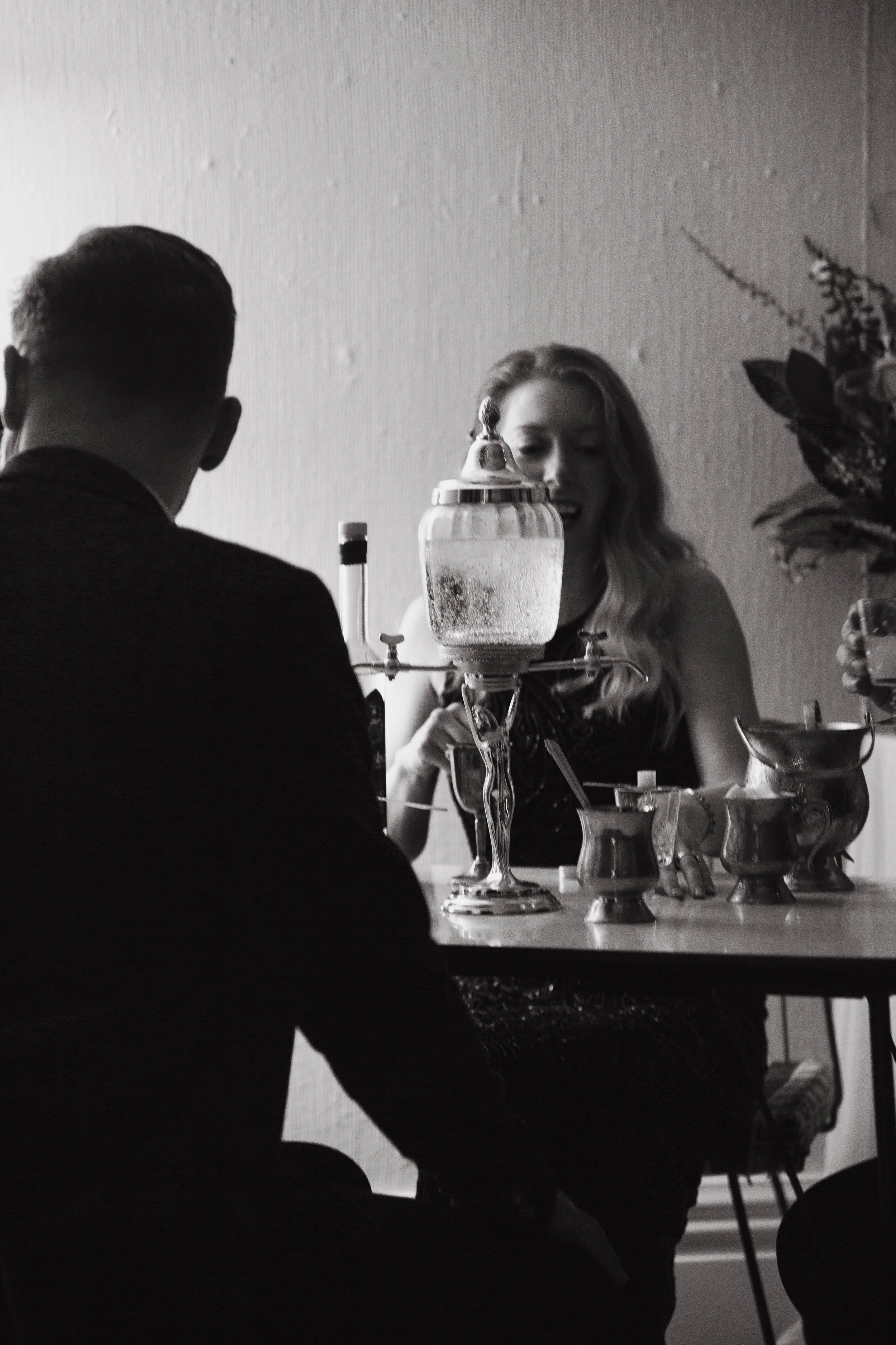 Black-and-white photo of two people playing a game of chess at a table. A glass container with a spout is in the center, and a plant is in the background.