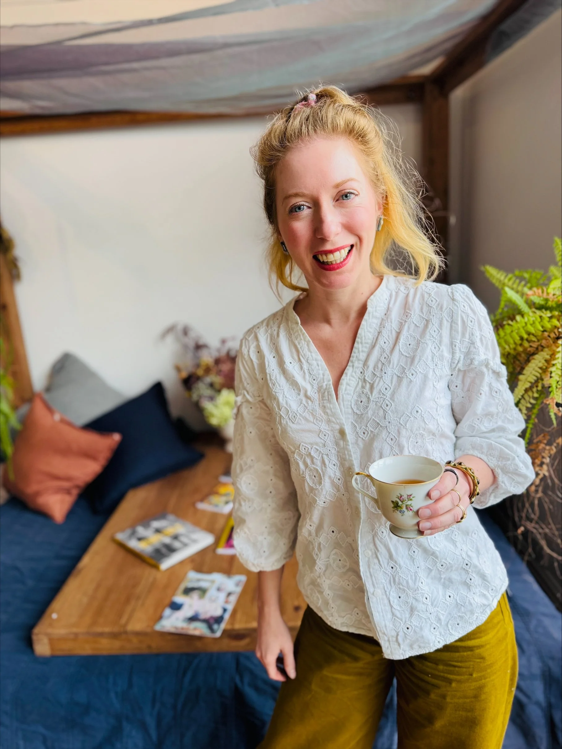 A woman with blonde hair in a bun, wearing a white embroidered shirt and mustard-yellow pants, is holding a tea cup and smiling. Behind her is a wooden table with scattered photographs, a blue couch with orange and blue pillows, and a plant.
