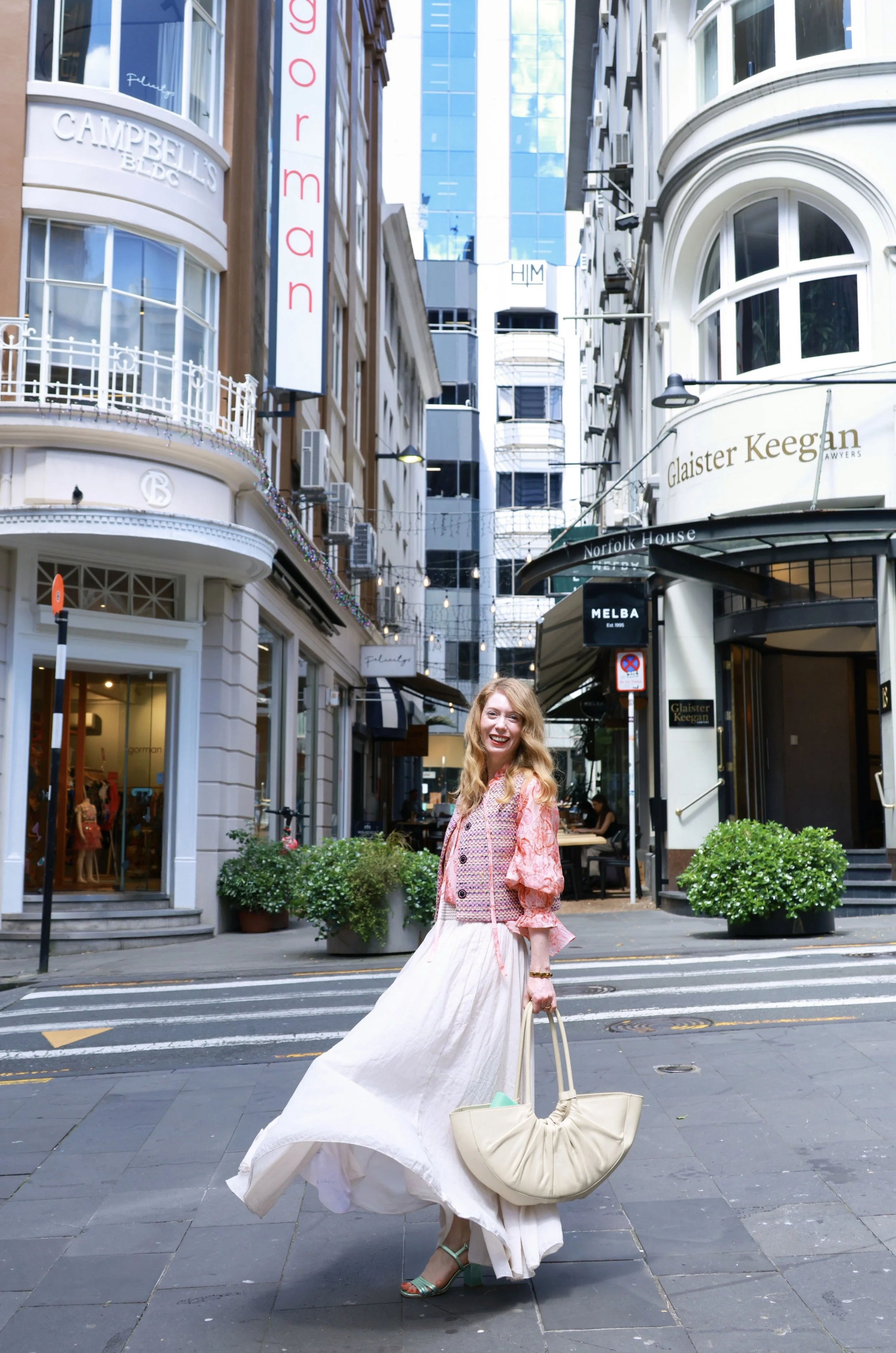 A woman dressed in a flowing white skirt, patterned pink and orange jacket, and green high heels is walking down a city street. She is carrying a large cream-colored handbag and smiling at the camera. The street is lined with shops and signs, including Glaister Keegan and Melba, with buildings featuring large windows and outdoor seating areas.