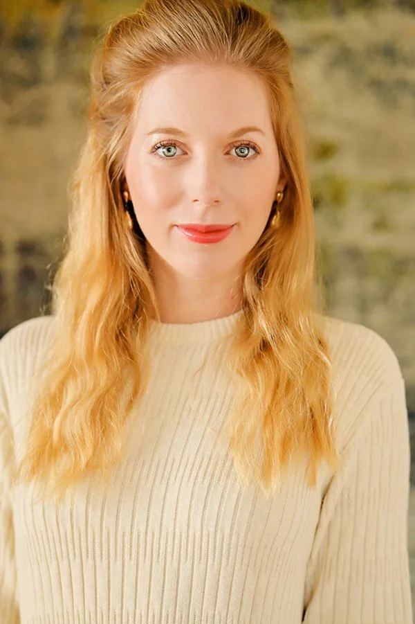 Portrait of a young woman with long, wavy blonde hair wearing a cream-colored ribbed sweater, standing outdoors with a blurred green and brown background.
