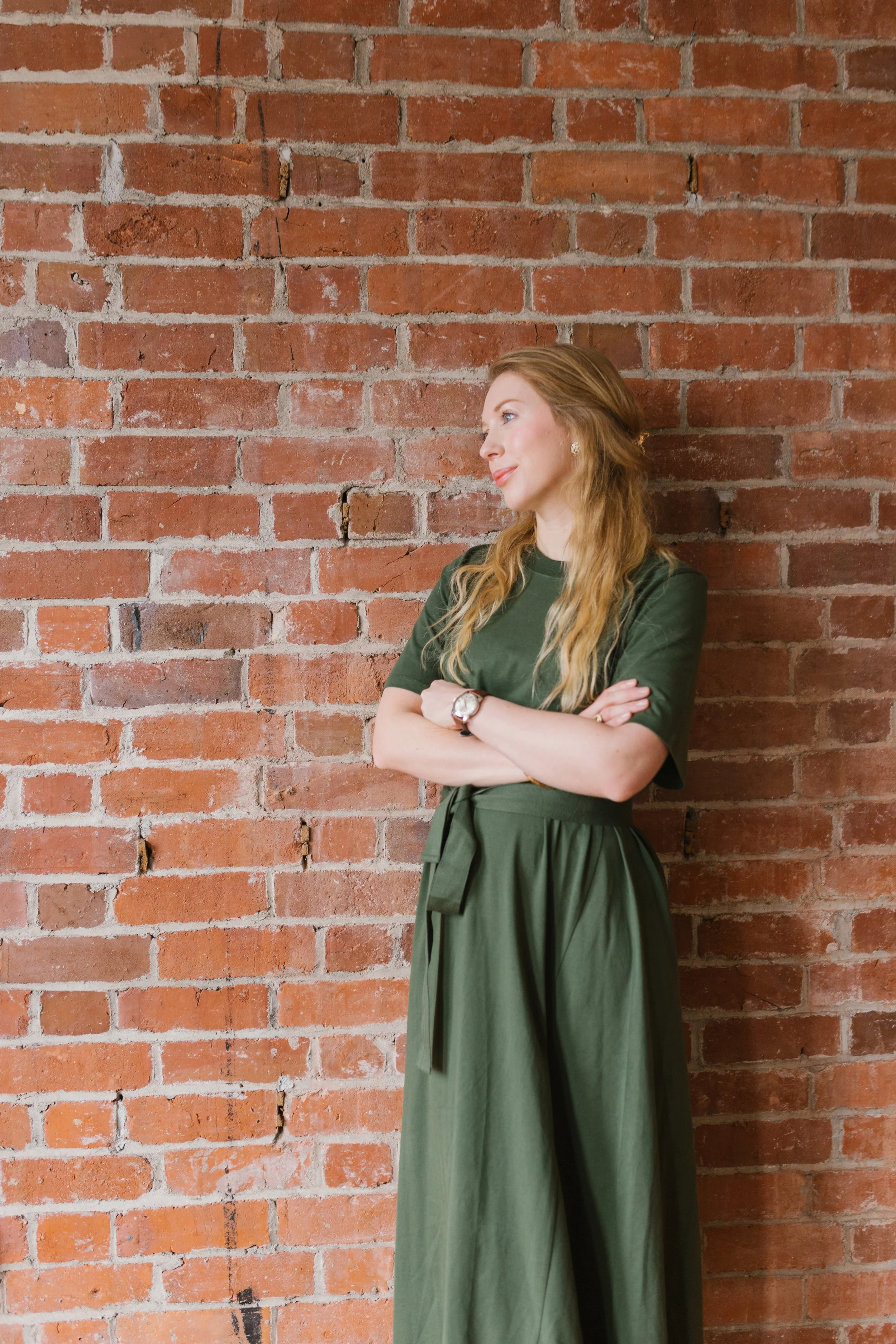 A woman with long, wavy red hair standing against a red brick wall. She is wearing a dark green dress with short sleeves, crossed arms, and a watch on her left wrist. She has a slight smile and is gazing sideways.