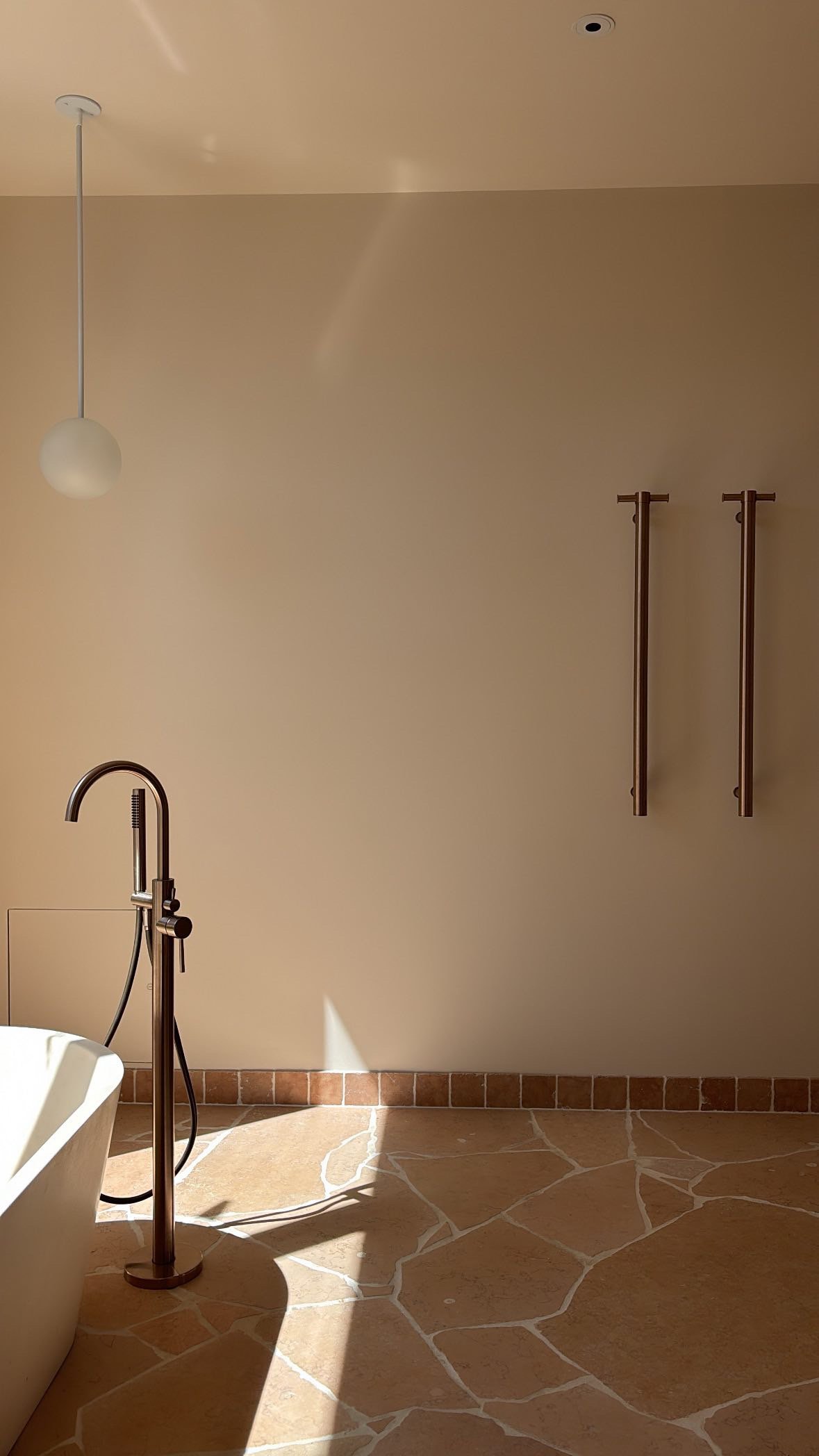A bathroom with a beige wall, a hanging white globe light, a modern black faucet, and a freestanding bath. There are two wall-mounted towel bars without towels and stone tile flooring.