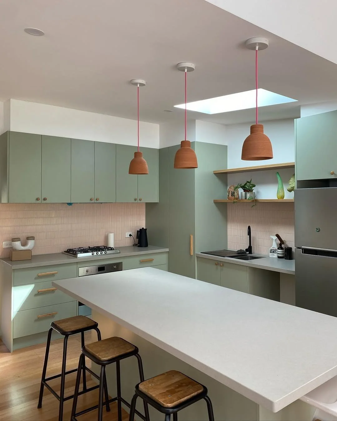 Modern kitchen with green cabinets, pink backsplash, and a white countertop island, featuring three terracotta pendant lights, open shelves with plants and decorative objects, black faucet, and appliances including a refrigerator and a stove.