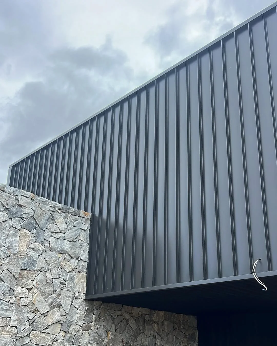 Close-up of the corner of a modern building with dark gray metal siding and a stone wall underneath, under an overcast sky.
