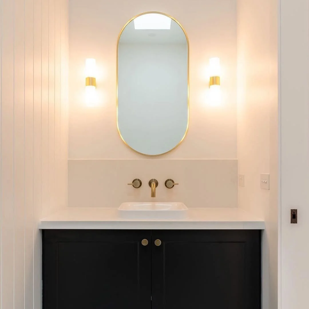 Modern bathroom vanity with an oval mirror framed in gold, two wall sconces, a white sink, and a black cabinet, with a white countertop.