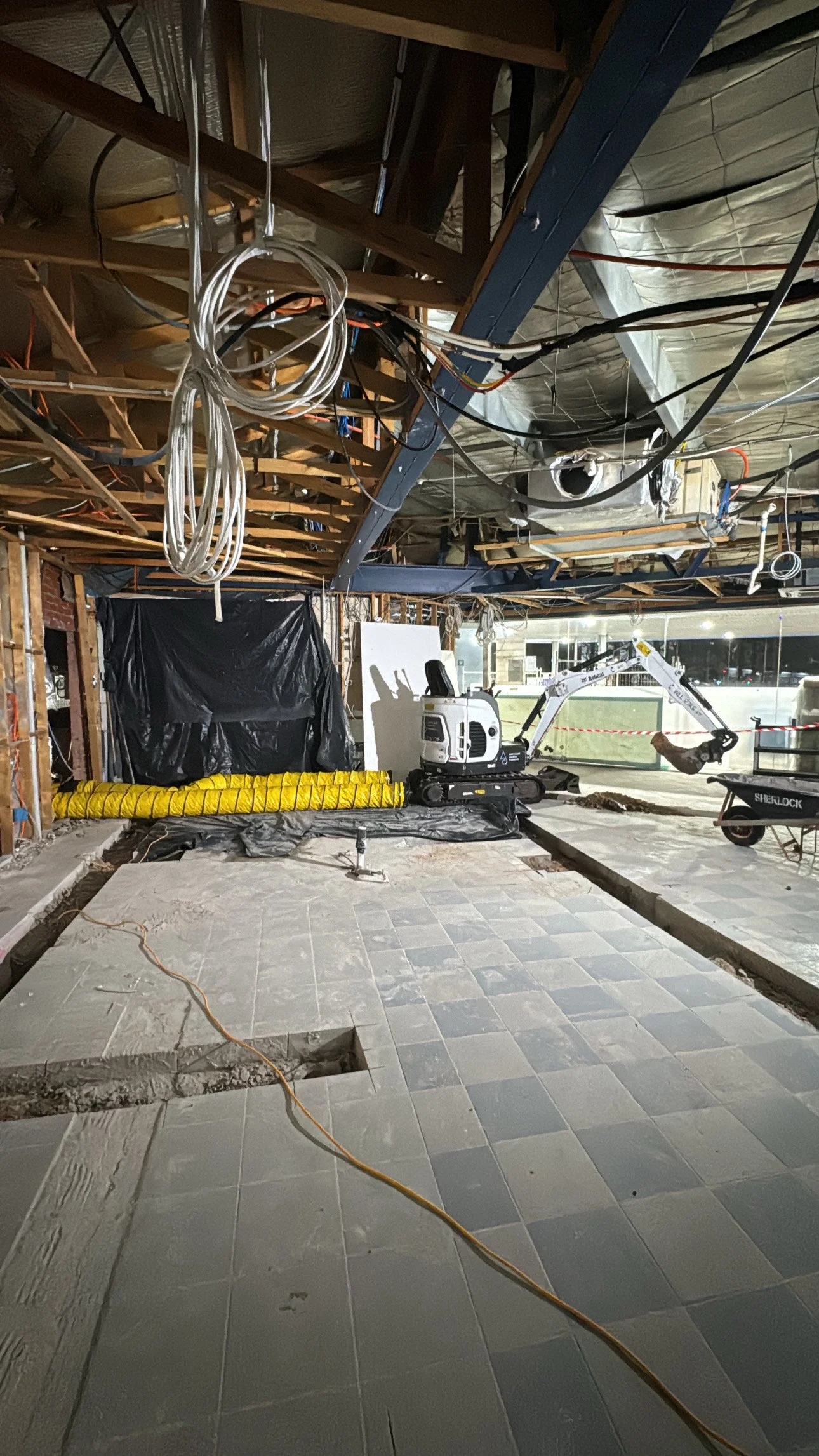 Construction site with exposed ceiling framework, electrical wiring, and a small excavator working on the floor. The area appears to be indoors with tiled flooring partially removed and construction tools around.
