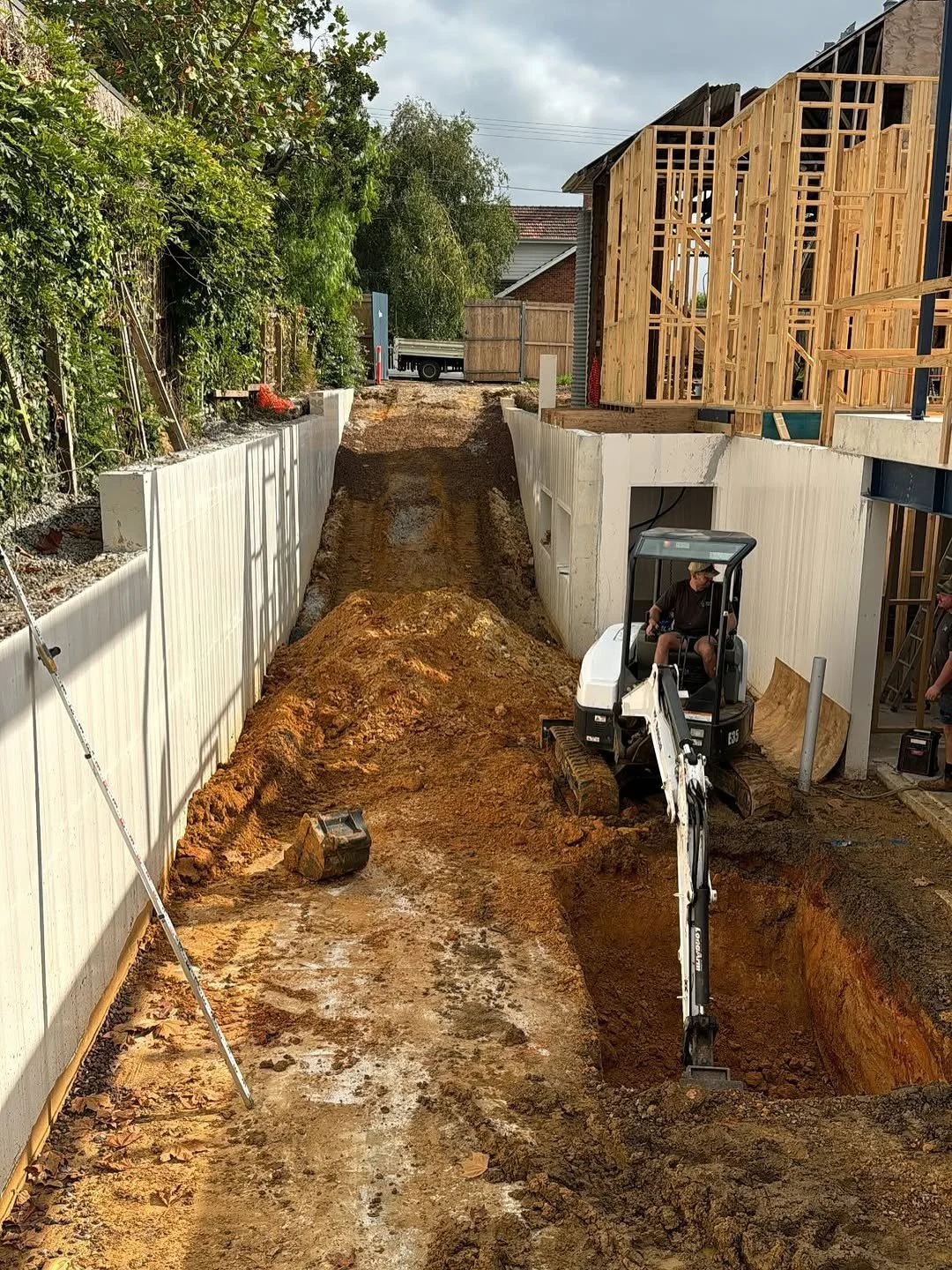 Construction site with a small excavator digging a trench, wooden framing of a building under construction, and a white retaining wall on both sides of the dirt path.