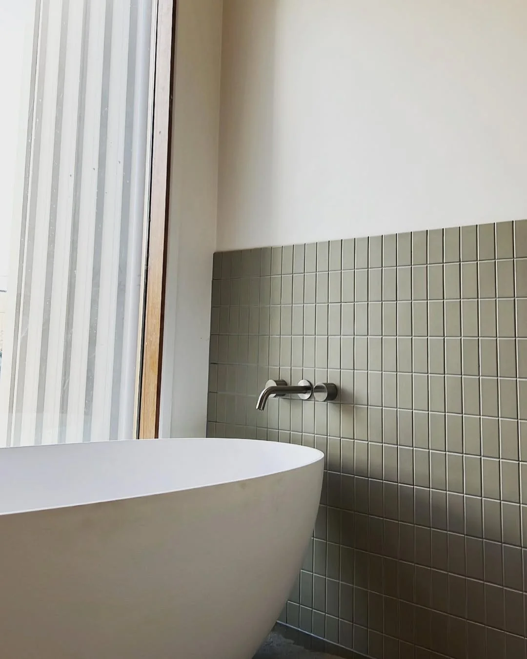 Modern bathroom with white free-standing bathtub, small square beige tiles on a half wall, a wall-mounted faucet, and a large window with a sliding door.
