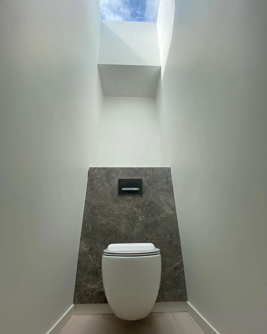 Modern toilet in a minimalist bathroom with a skylight ceiling, white walls, and gray marble wall behind the toilet.