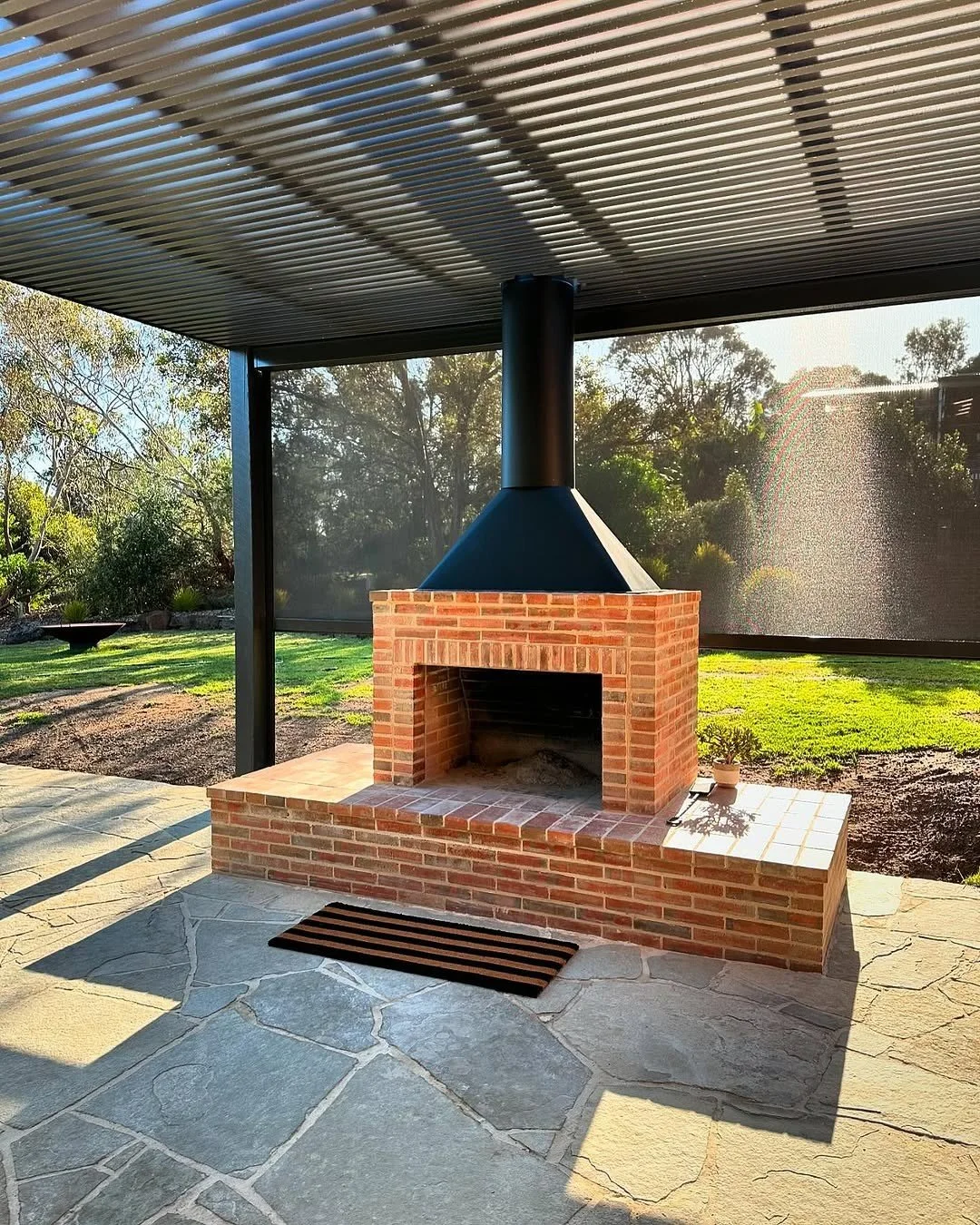 Exterior view of a brick outdoor fireplace with a black chimney, set on a stone patio under a metal roof, with green yard and trees in the background.