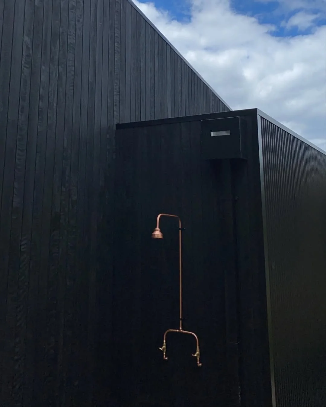 Dark modern premium cladding house with vertical paneling, an outdoor shower with copper piping and a copper showerhead, against a cloudy sky.