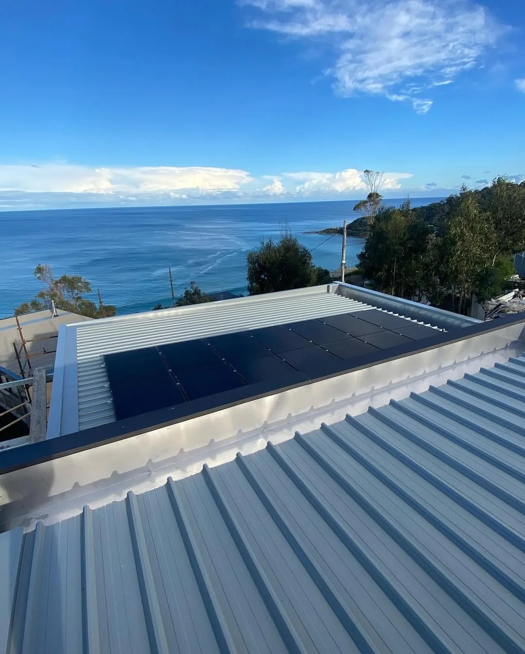 View of surf coast ocean from a rooftop with solar panels installed on corrugated metal roofing, trees, and distant land under a partly cloudy sky.