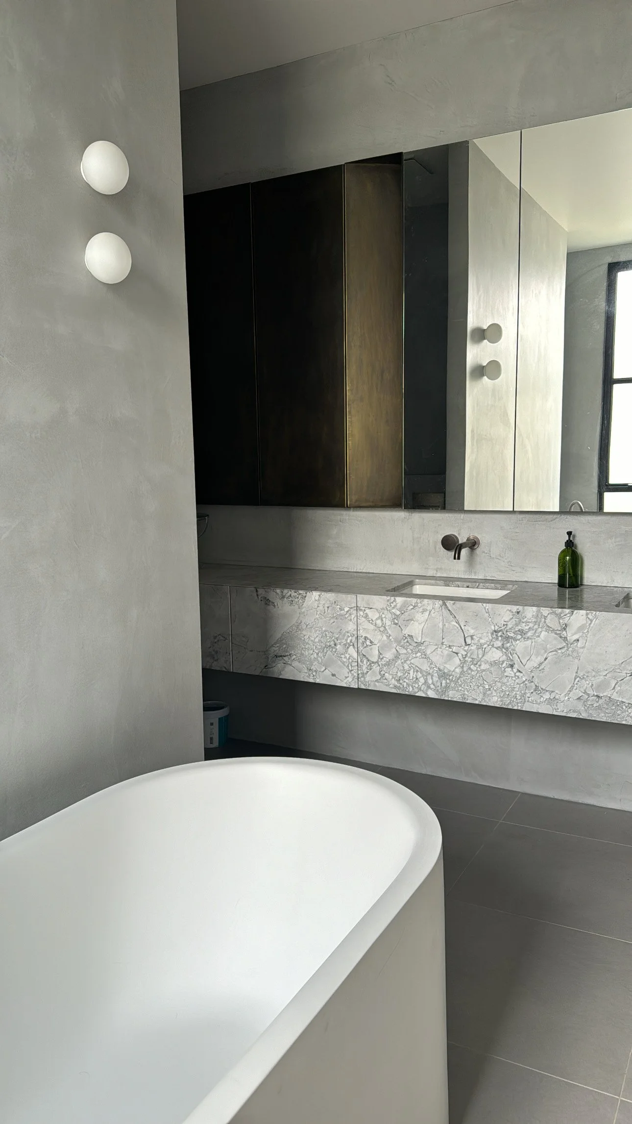 Modern bathroom with concrete walls, a freestanding white bathtub, marble vanity with integrated sink, large mirror, wall-mounted faucet, and a window letting in natural light.