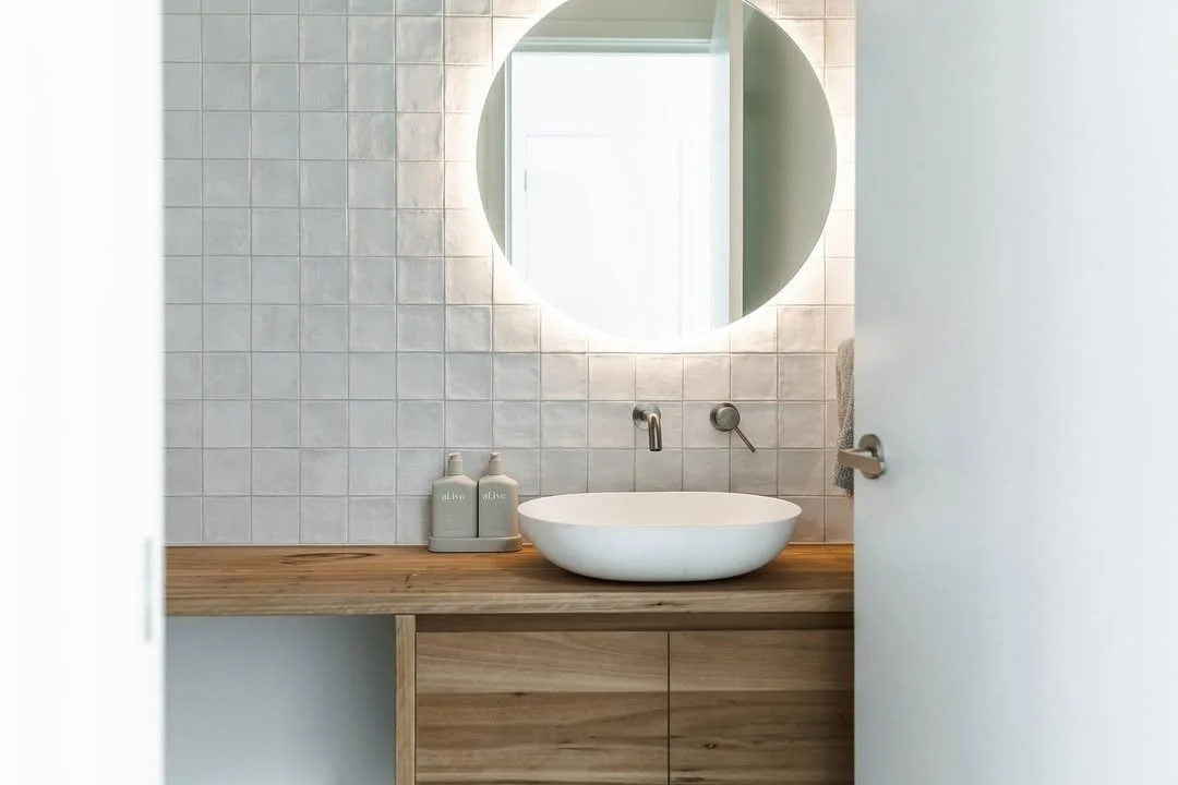 Bathroom with a round mirror, wall-mounted faucet, wooden vanity, vessel sink, and soap dispensers.