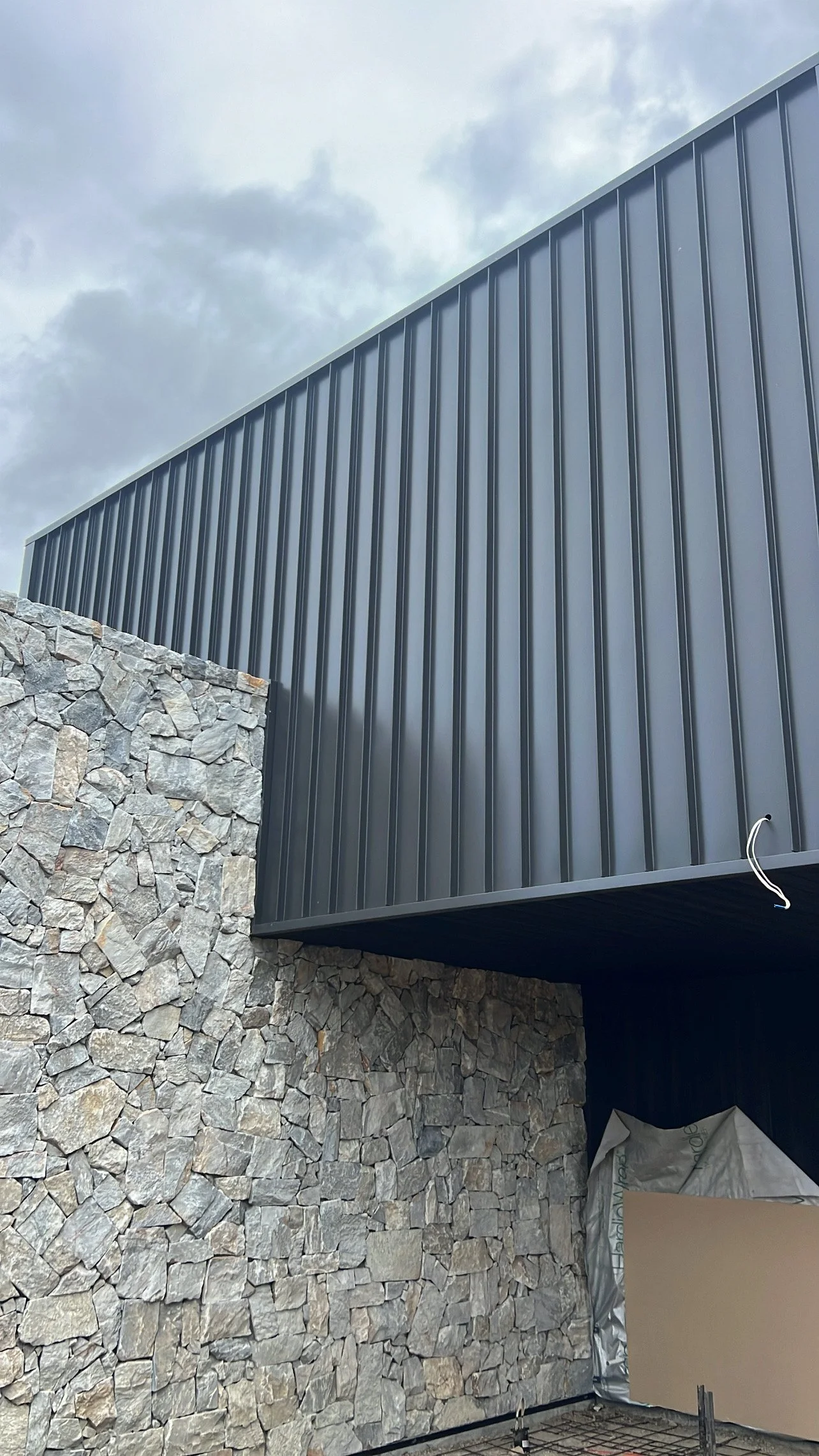 A modern building exterior featuring a stone wall and a sleek, dark metal overhang, with a partly cloudy sky in the background.
