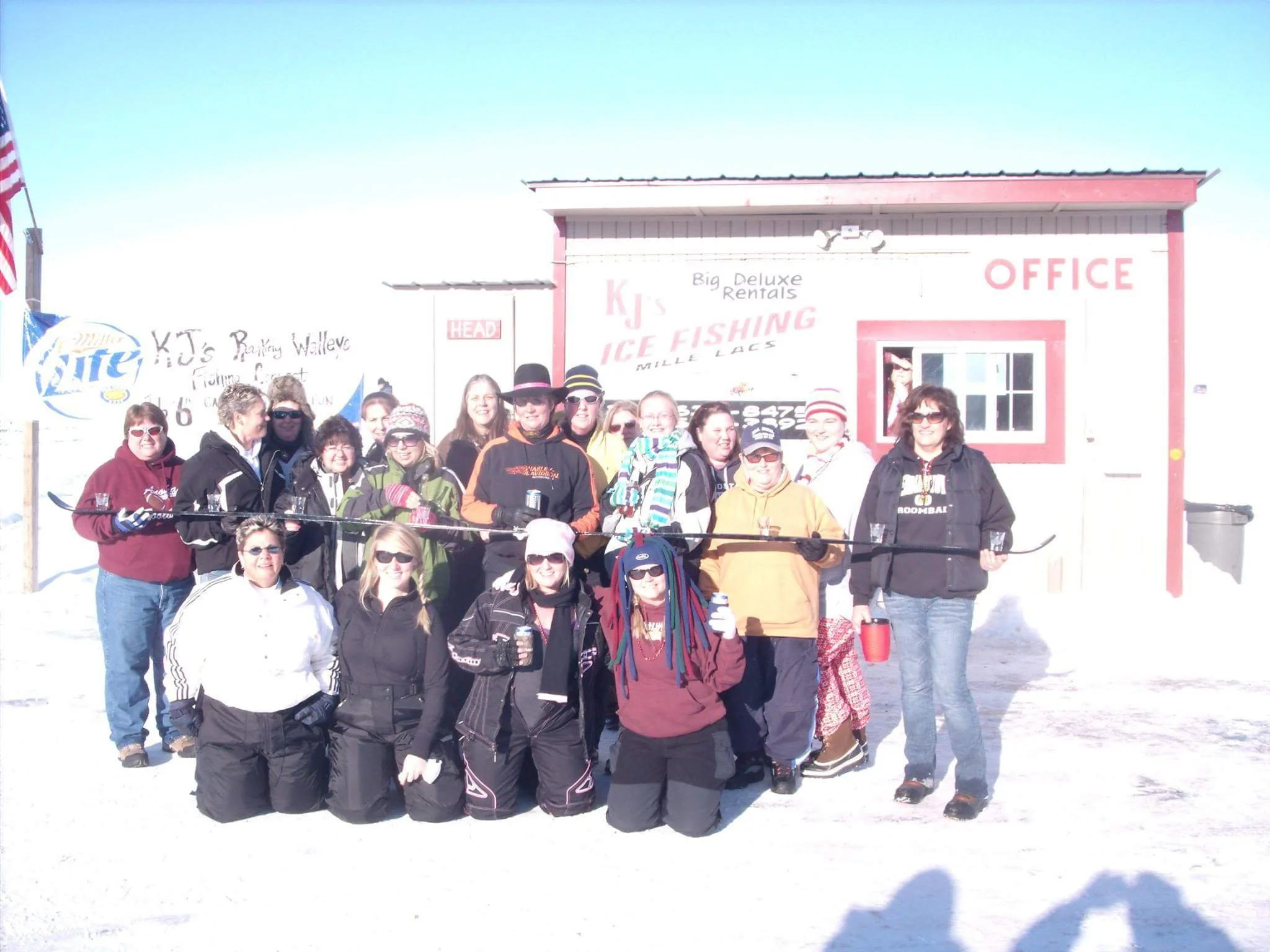 Group of people in winter clothing standing on snow in front of a building with signs that say 'Ice Fishing' and 'Office.' They are holding hockey sticks and wearing sunglasses.