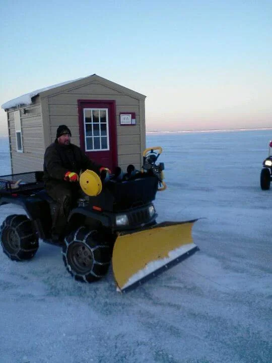 Person riding a snowmobile with a yellow snow plow attachment on a snowy, flat landscape near a small beige shed with a red door and window, and a partly cloudy sky in the background.