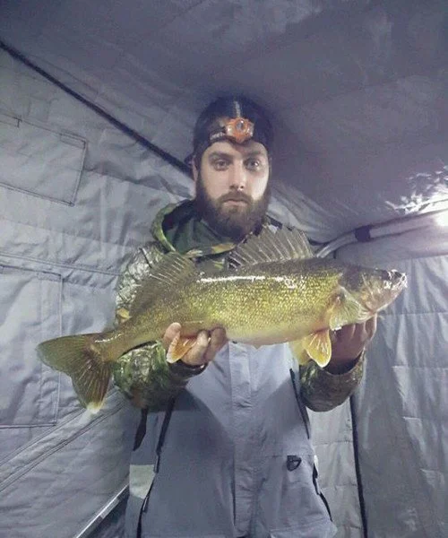 Man wearing a headlamp holding a large fish inside a tent.