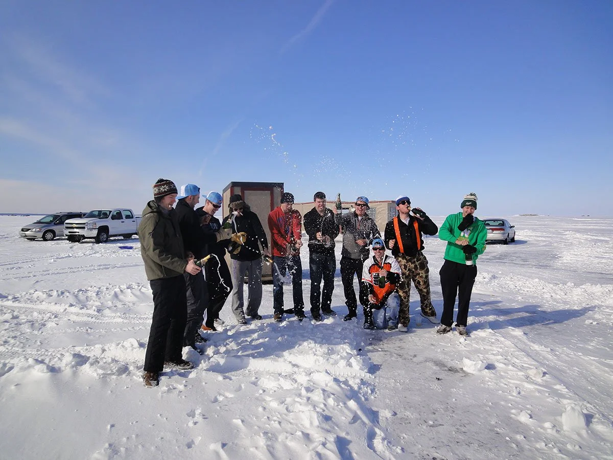 Group of people celebrating outdoors in snowy landscape, popping champagne, and dressed warmly in winter clothing, with cars in the background.