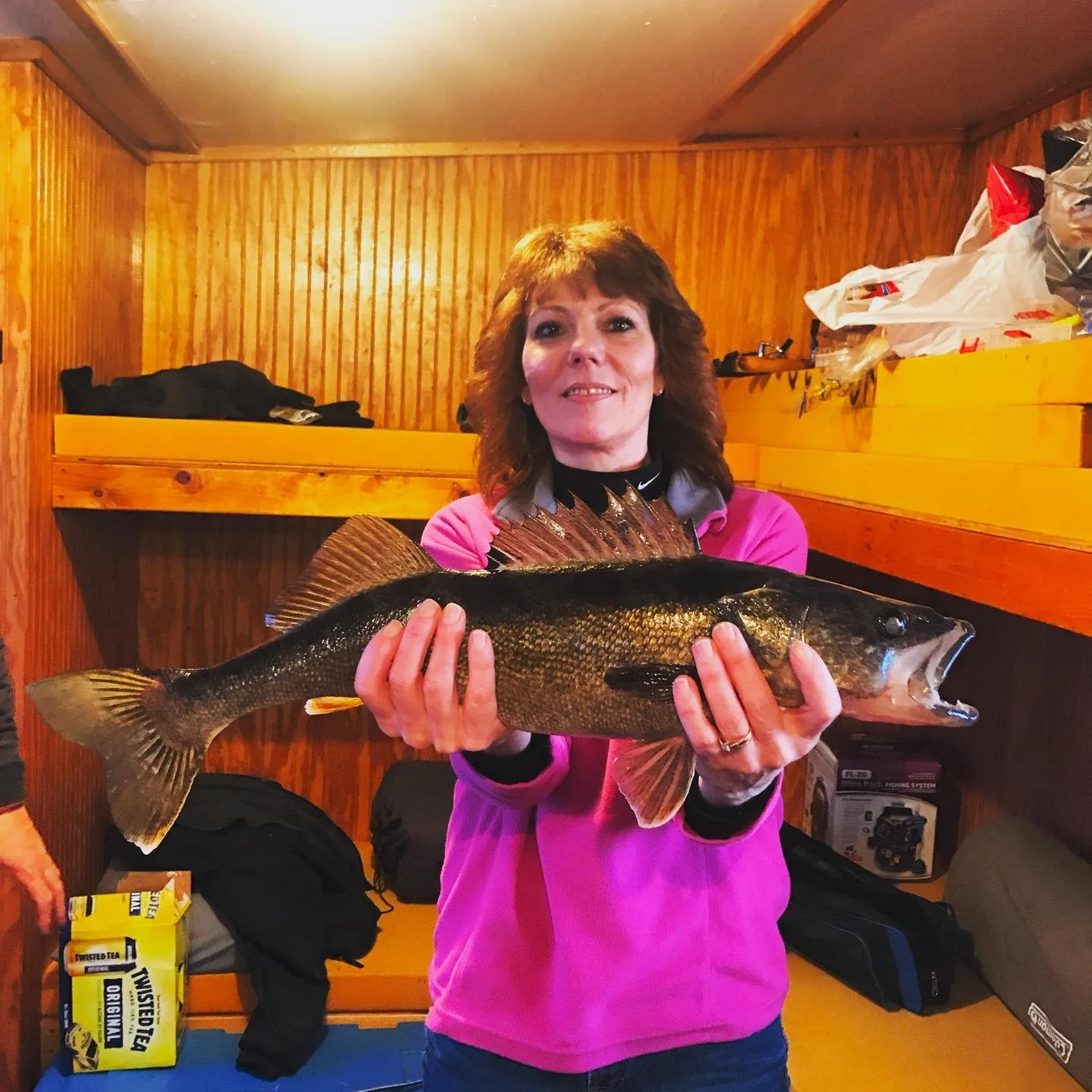 Woman holding a large fish indoors with wood-paneled walls, wearing a pink jacket and smiling.