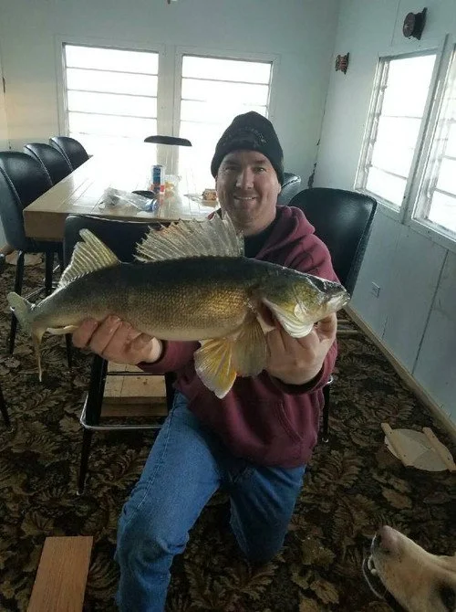 Man wearing black hat and maroon hoodie sitting indoors, holding a large fish with a greenish top and light belly.