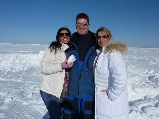 Three people standing close together in a snowy landscape, smiling at the camera, with one holding a snowball and dressed in winter clothing.
