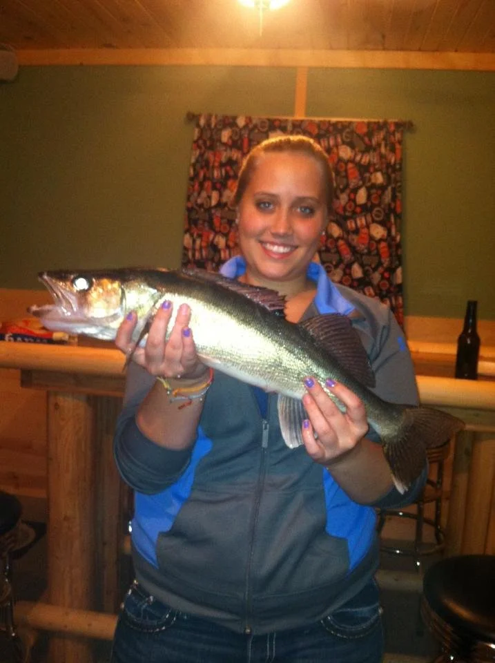 Woman smiling and holding a large fish inside a cozy room with wood-paneled ceiling and green walls.