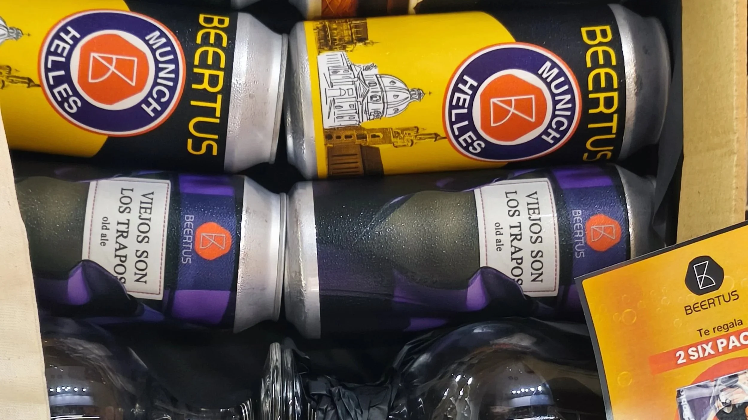 Latas de cerveza Beertus con etiquetas que dicen "Viejitos Son Los Trapos Old Ale" y cartel de promoción de un paquete de 2 latas en una caja