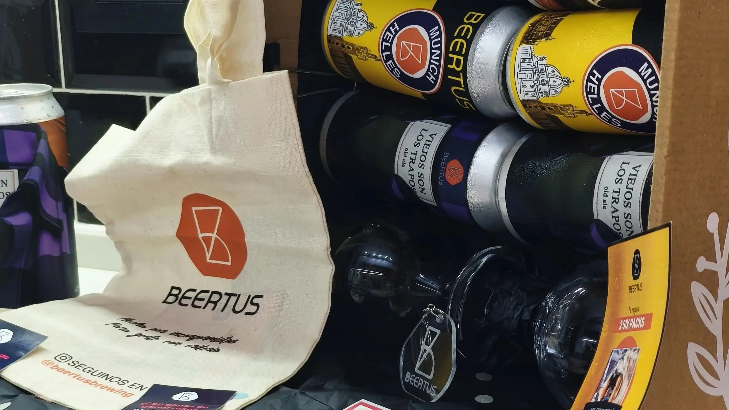 Latas de cerveza Beertus, una bolsa de tela con el logo de Beertus, un vaso de cristal vacío con etiquetas de Beertus, y una caja promocional de 2 packs de cerveza Beertus sobre una superficie de madera.