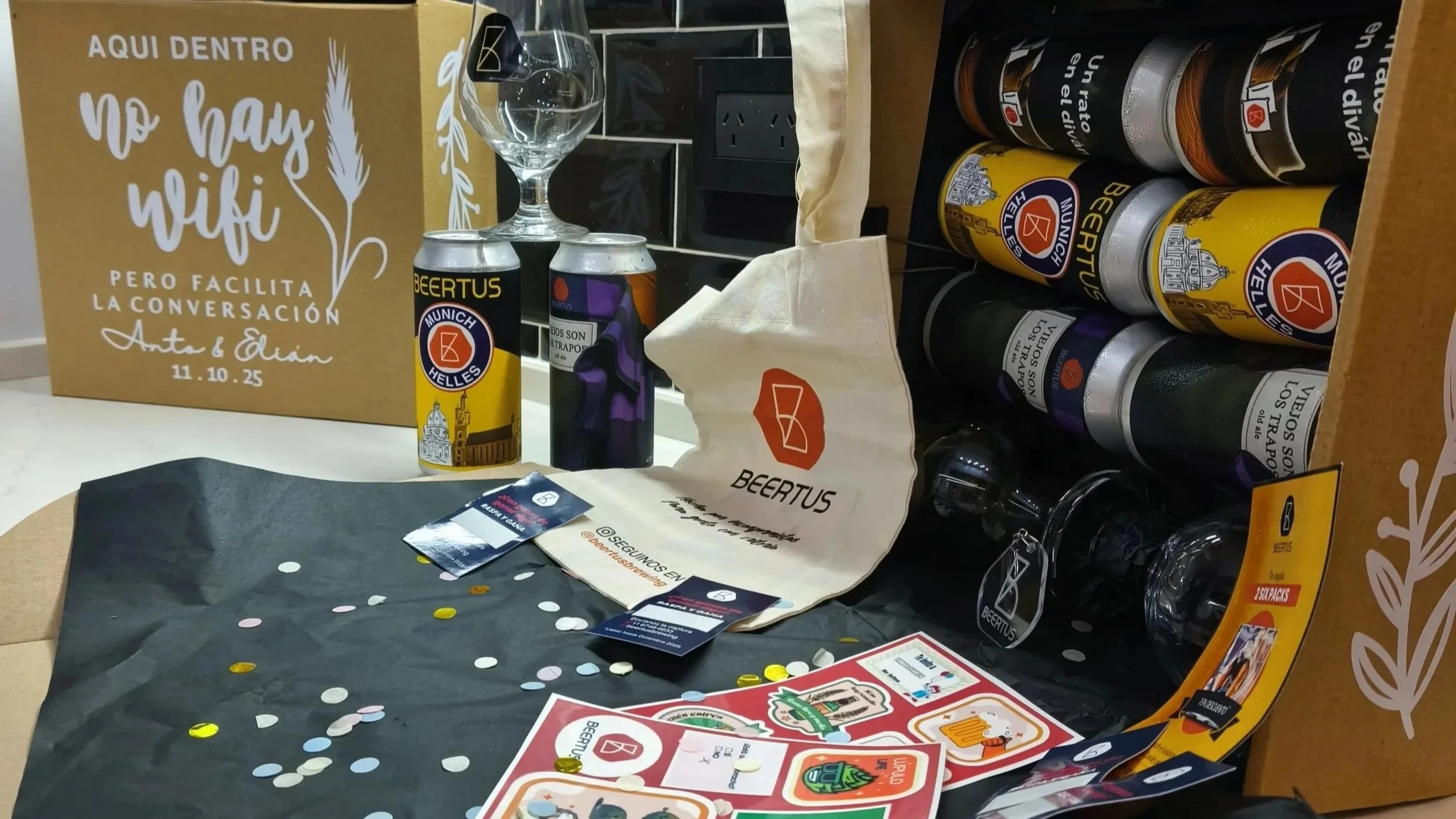 Un conjunto de artículos promocionales de Beertus en una mesa, incluyendo latas de cerveza, un vaso, etiquetas, confeti y una caja grande con un cartel en el fondo que dice 'Aquí dentro no hay wifi, pero facilita la conversación'.