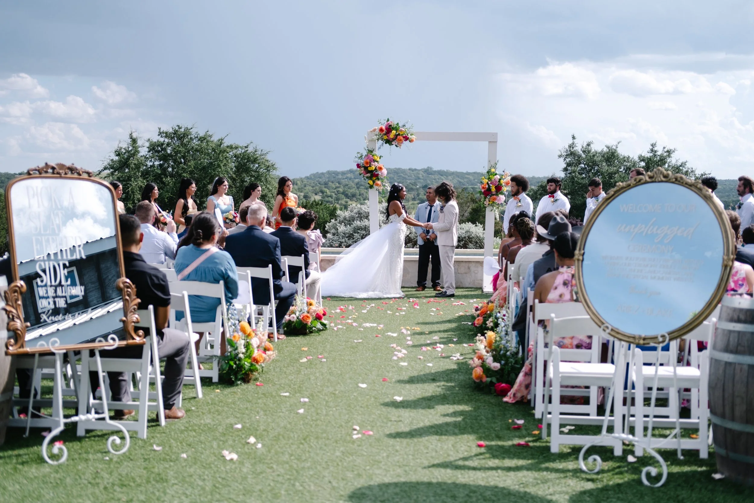 A scenic outdoor wedding ceremony with a bride and groom exchanging vows under a white arch decorated with colorful flowers, surrounded by guests seated on white chairs on a grassy area, with a scenic landscape and partly cloudy sky in the background