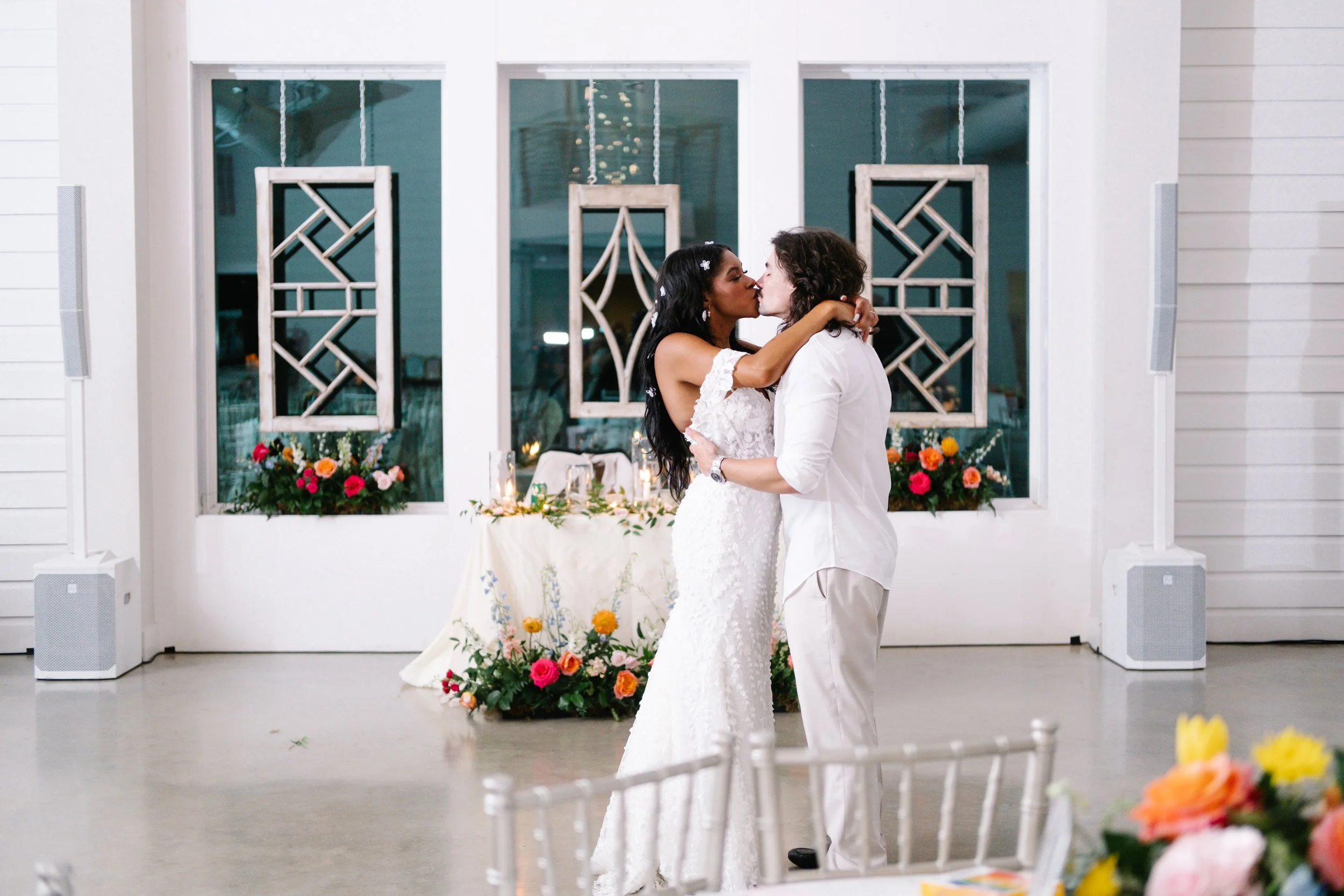 A bride and groom sharing a kiss during their wedding reception, with a decorated table and floral arrangements in the background.