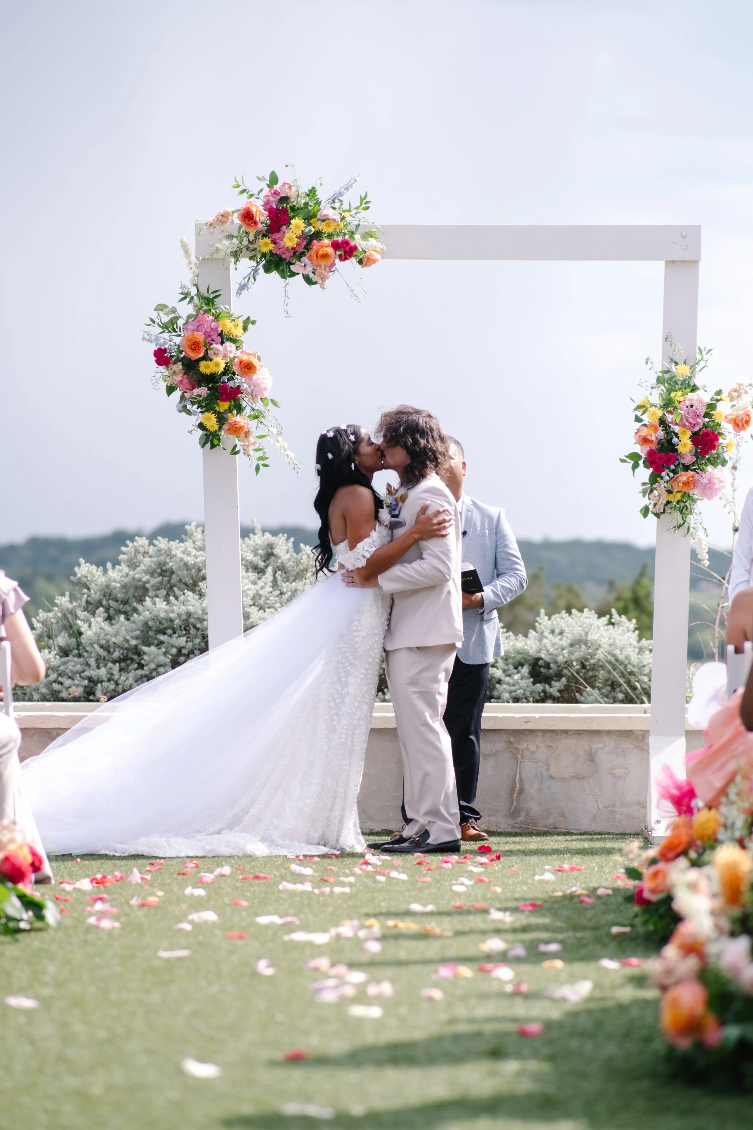 A same-sex couple sharing a kiss during their outdoor wedding ceremony under a floral arch, with guests and a scenic landscape in the background.