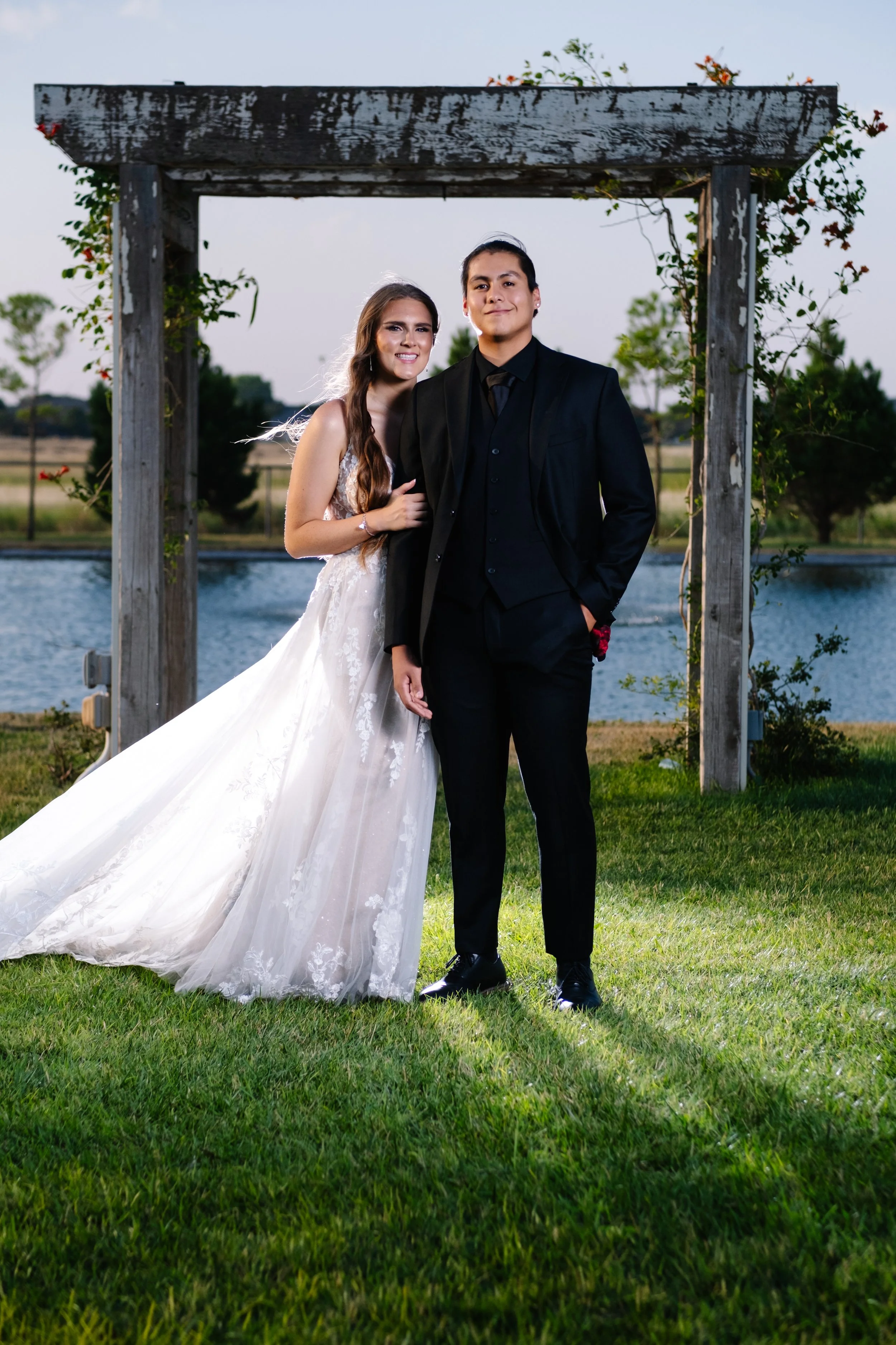 A bride and groom standing outdoors by a lake under a wooden archway, with trees and grass in the background, during their wedding celebration.