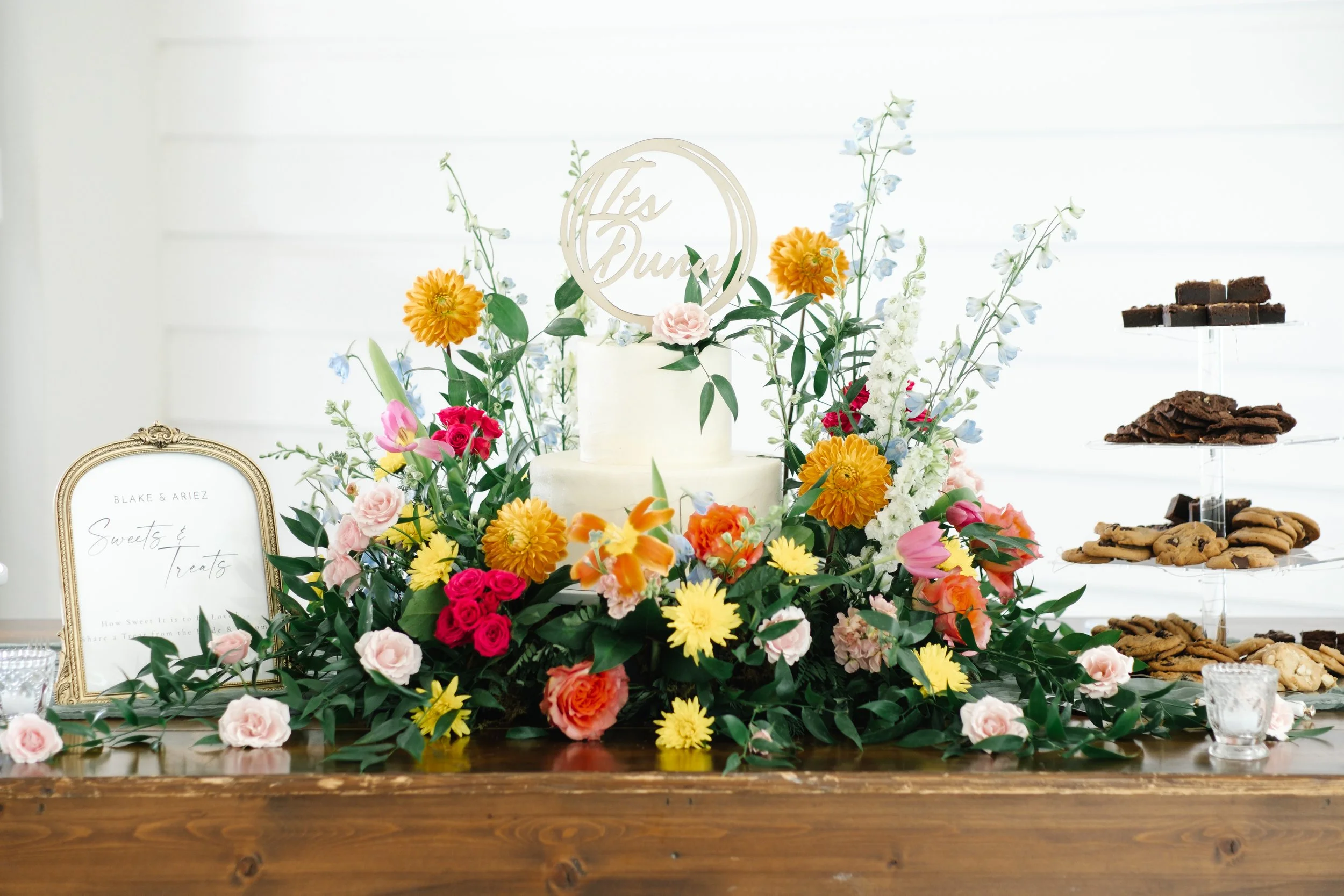 A wedding cake surrounded by colorful flowers, with a tray of cookies and a framed sign on a wooden table.