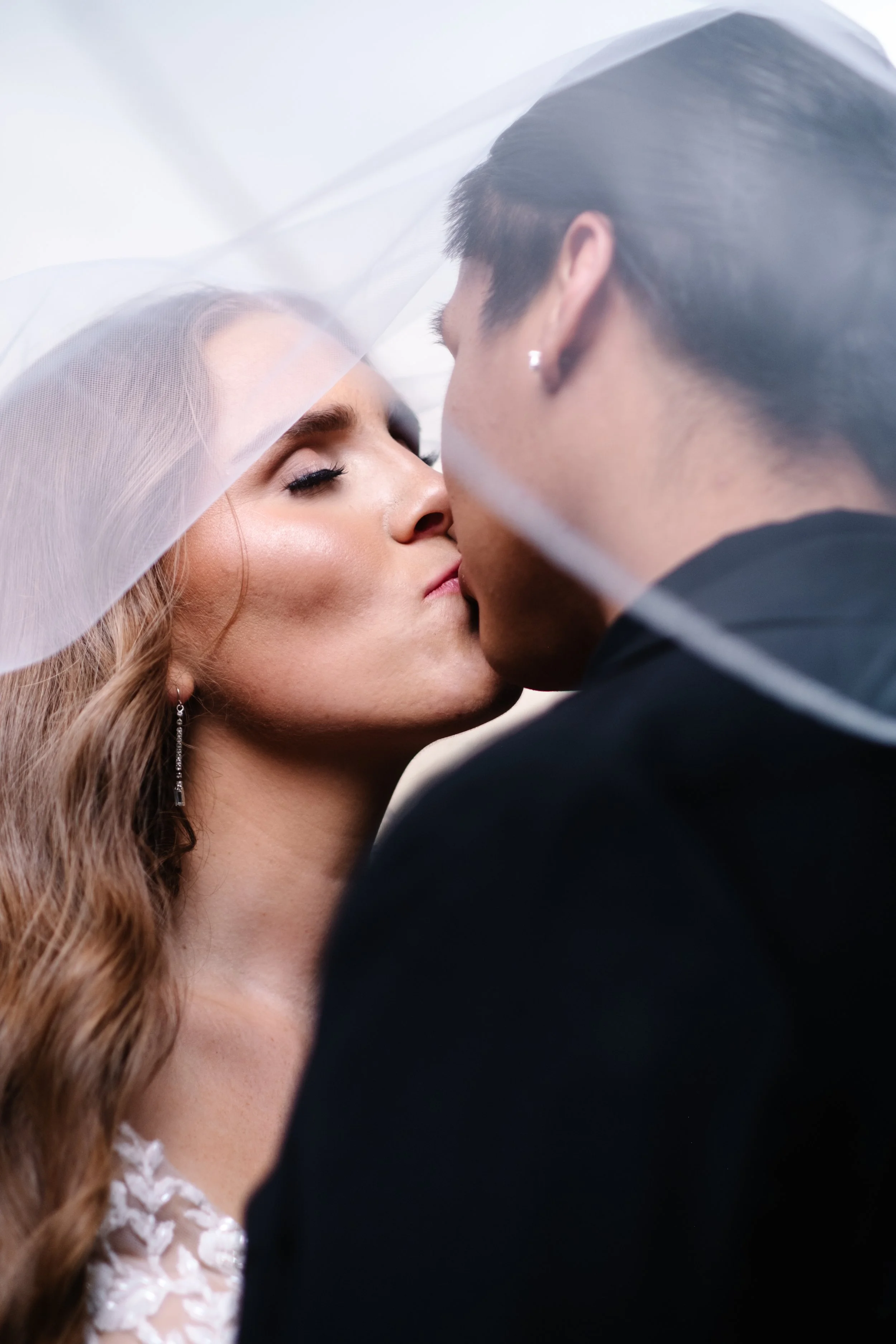 A bride and groom kissing under a sheer veil on their wedding day.