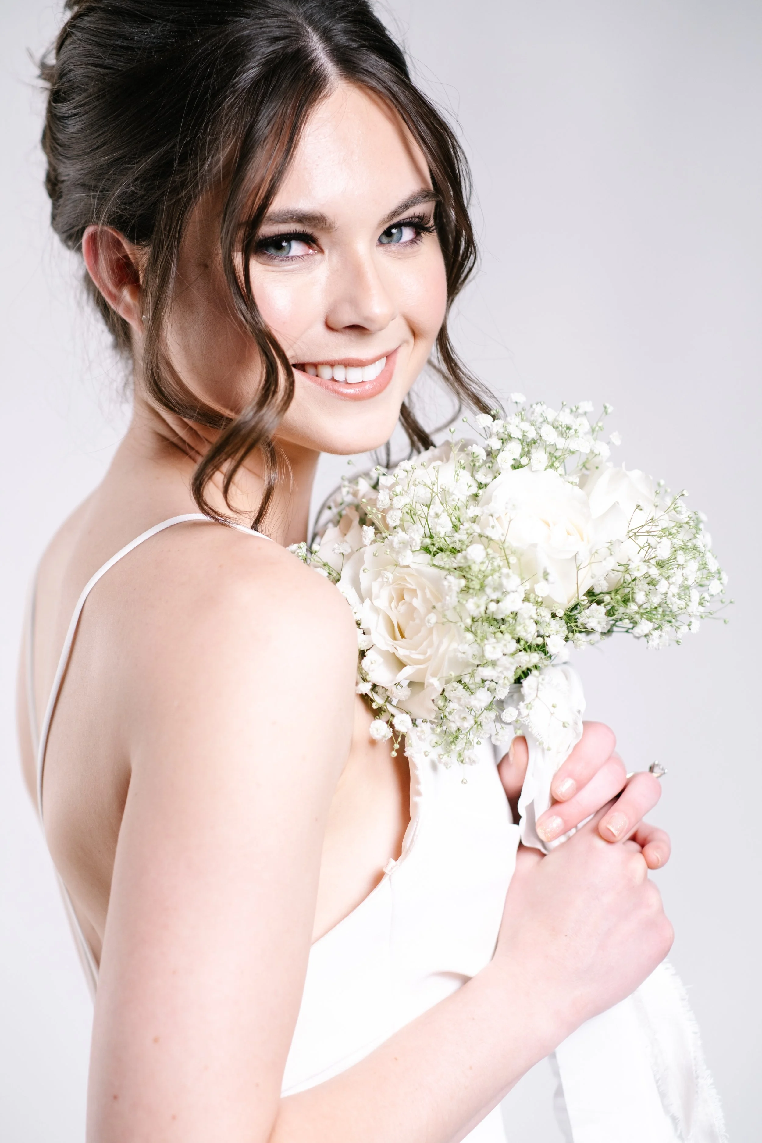 Bride with brown hair in loose curls holding a white bouquet of roses and baby's breath, smiling at camera.
