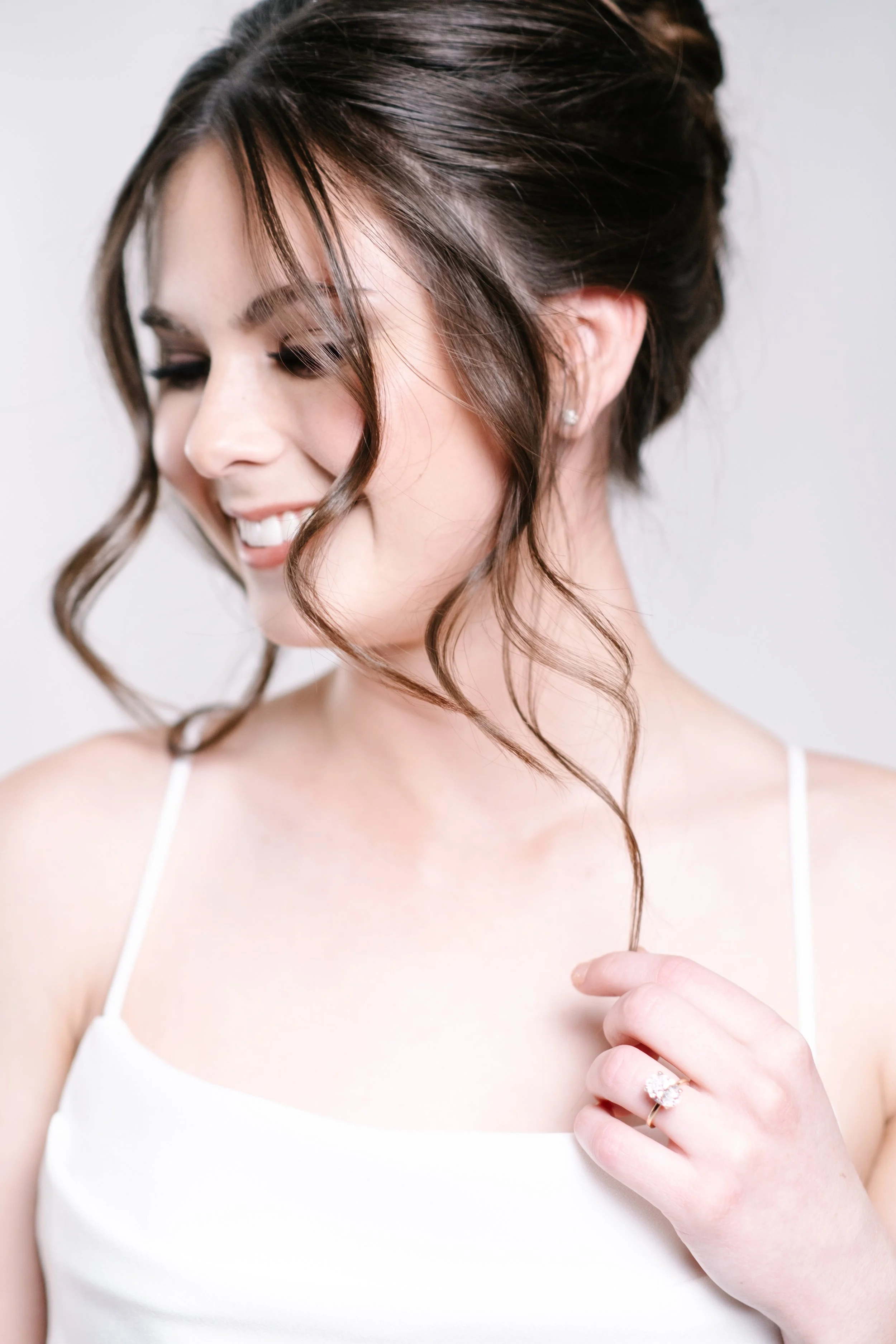 A woman with styled brown hair and a ring on her finger, wearing a white sleeveless top, smiling and looking down.