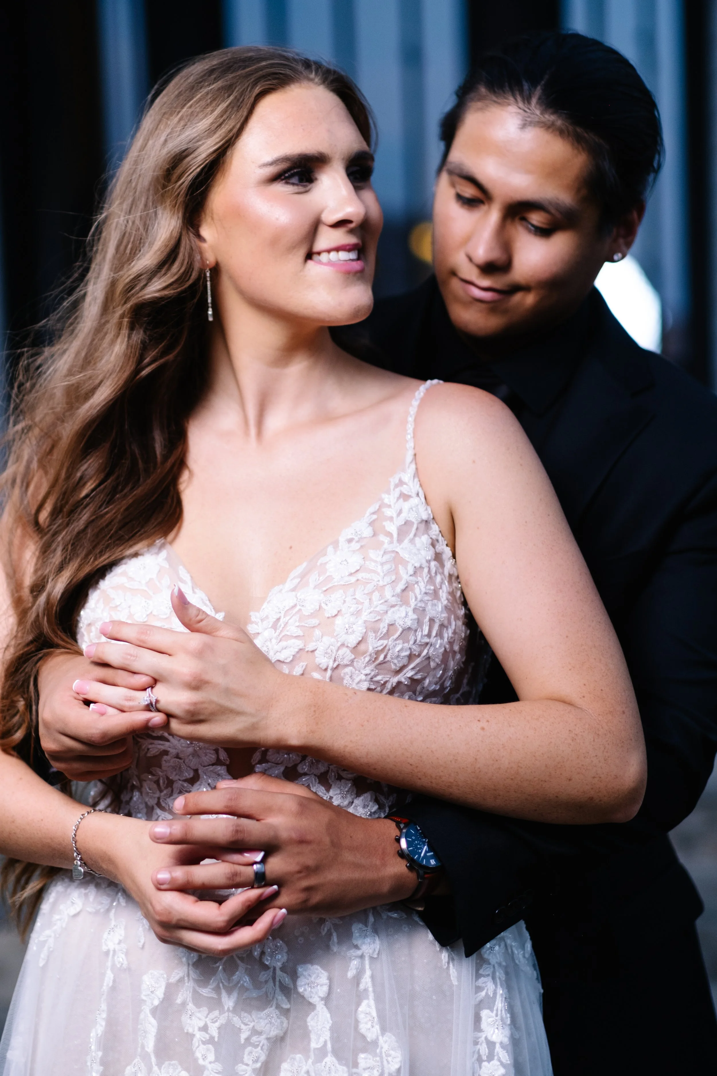 A couple dressed in formal attire embraces each other, with the woman wearing a lace wedding dress and the man in a black suit, in an intimate moment.
