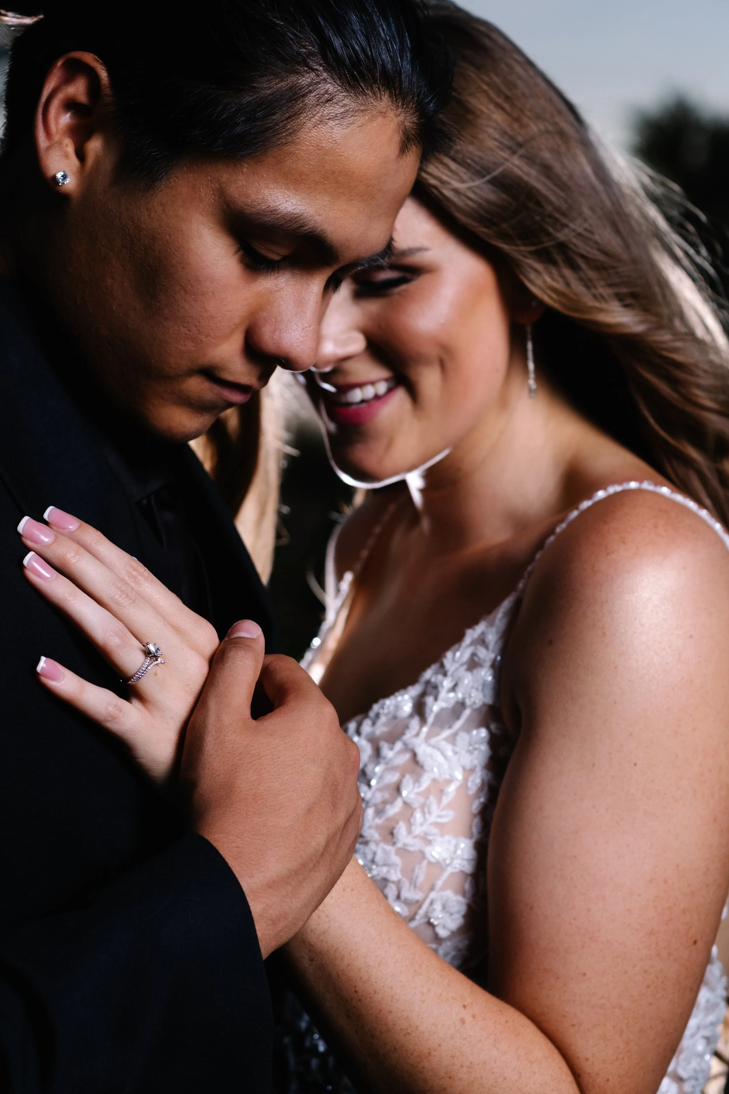 A close-up of a newlywed couple, with their foreheads touching and smiling, outdoors during sunset. The bride is wearing a lace wedding dress and the groom a black suit; the bride has an engagement ring on her finger.
