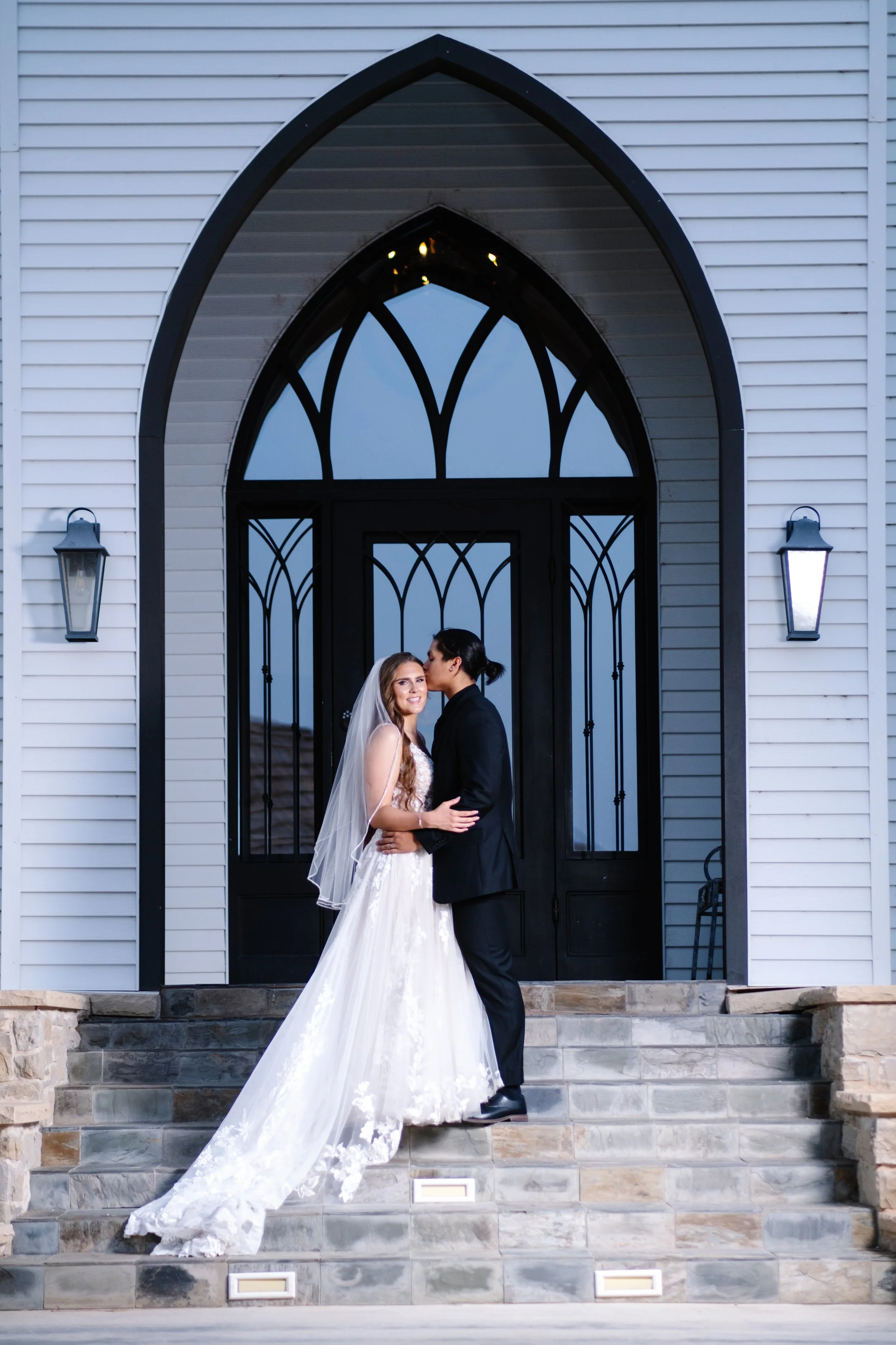 A bride and groom standing on the steps of a church, sharing a kiss, with the bride in a white wedding gown and veil, and the groom in a black suit, during their wedding celebration.
