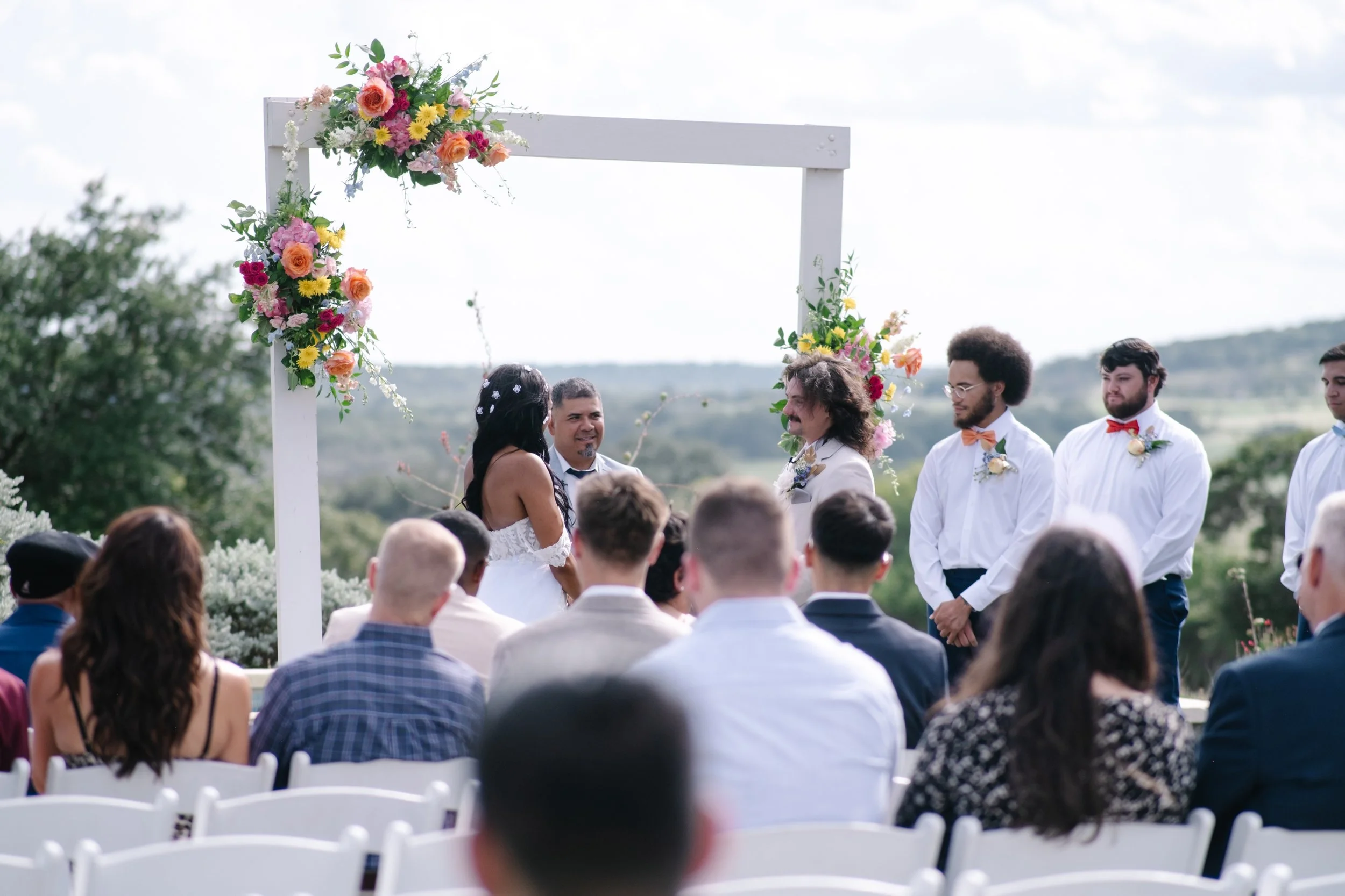 Outdoor wedding ceremony with a couple exchanging vows under a decorated arch, surrounded by guests, with a scenic landscape in the background.