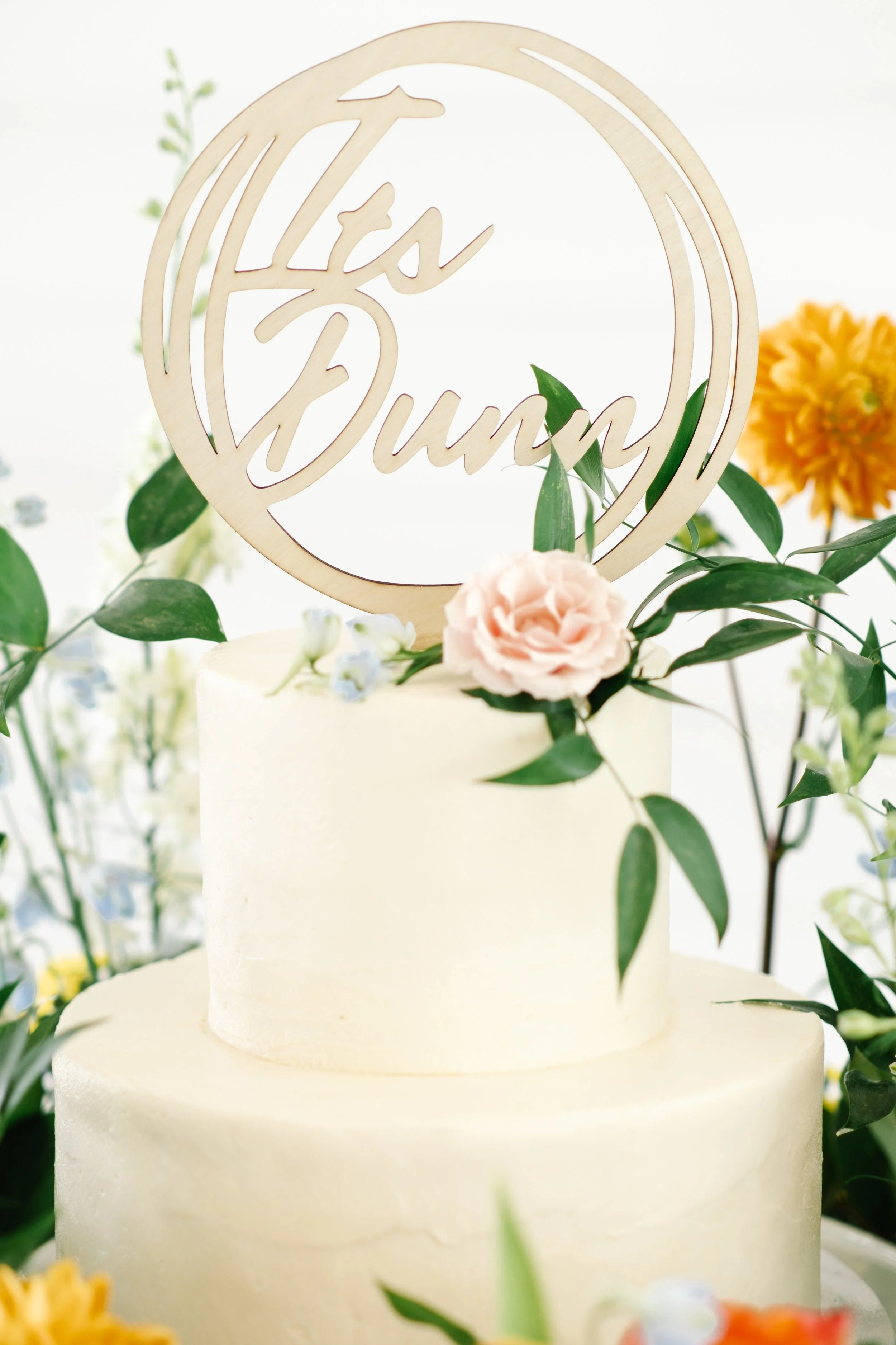 A wedding cake decorated with fresh flowers and a wooden cake topper that says 'Its Bunnies'.