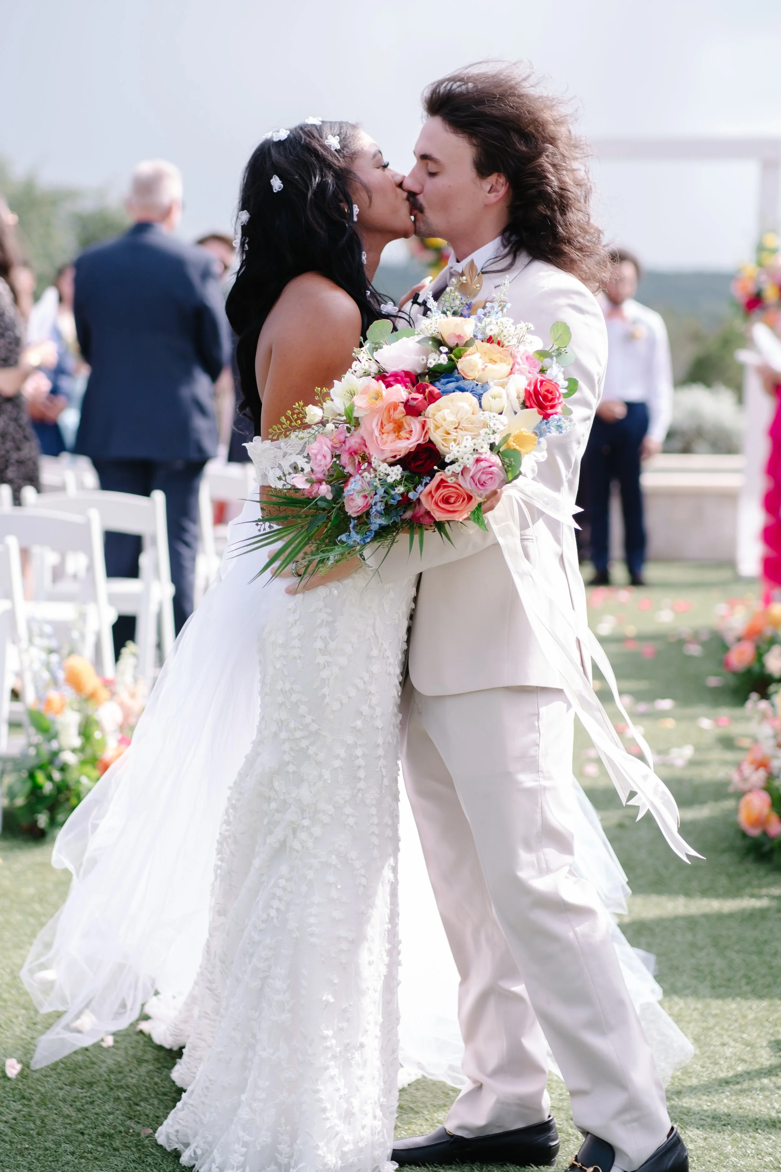 A bride and groom kiss during their wedding ceremony outdoors, with the bride holding a large bouquet of colorful flowers.