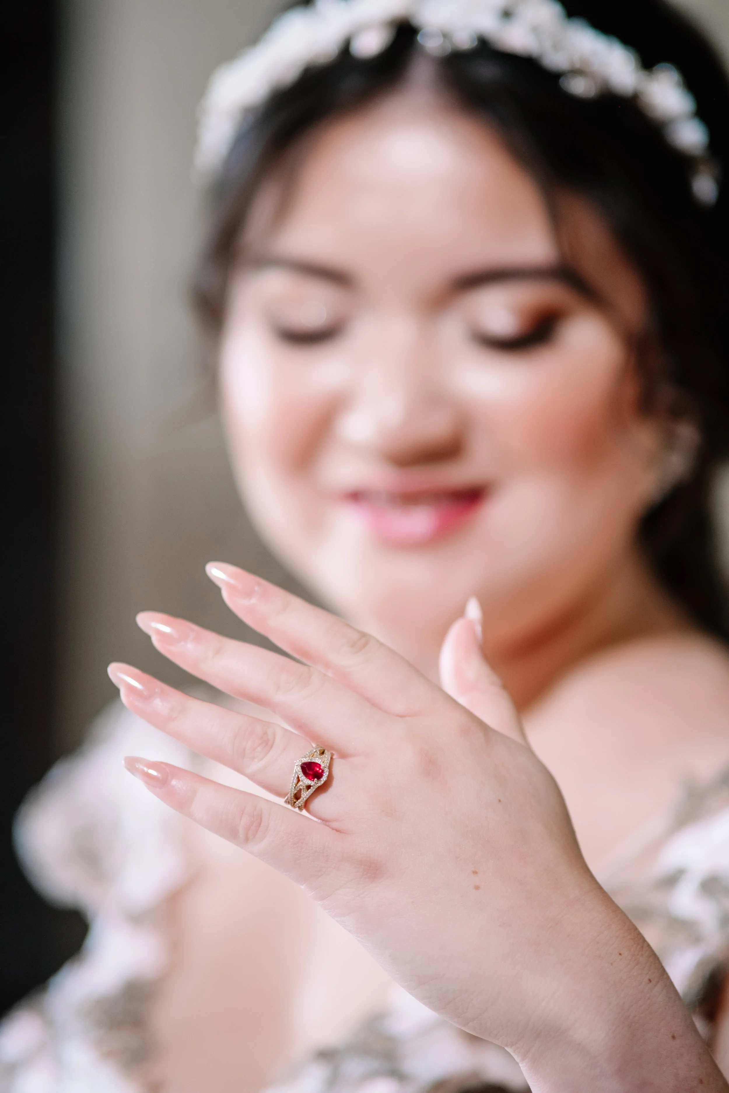 A woman shows off a ring with a red gemstone on her finger, smiling with her eyes closed and a blurred background.