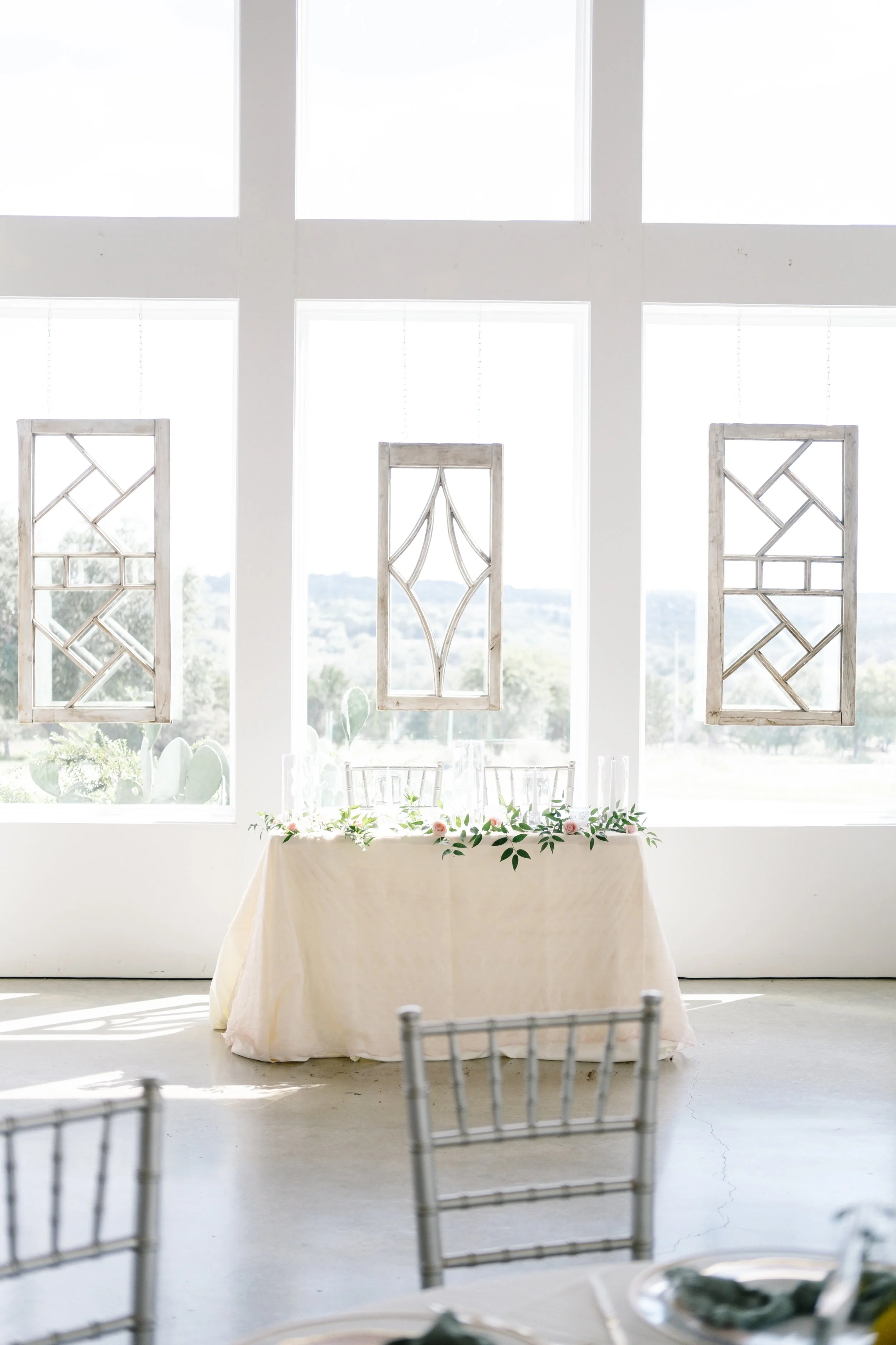 Wedding reception setup with a table covered in a cream tablecloth and floral greenery, positioned in front of large windows with decorative hanging panels.