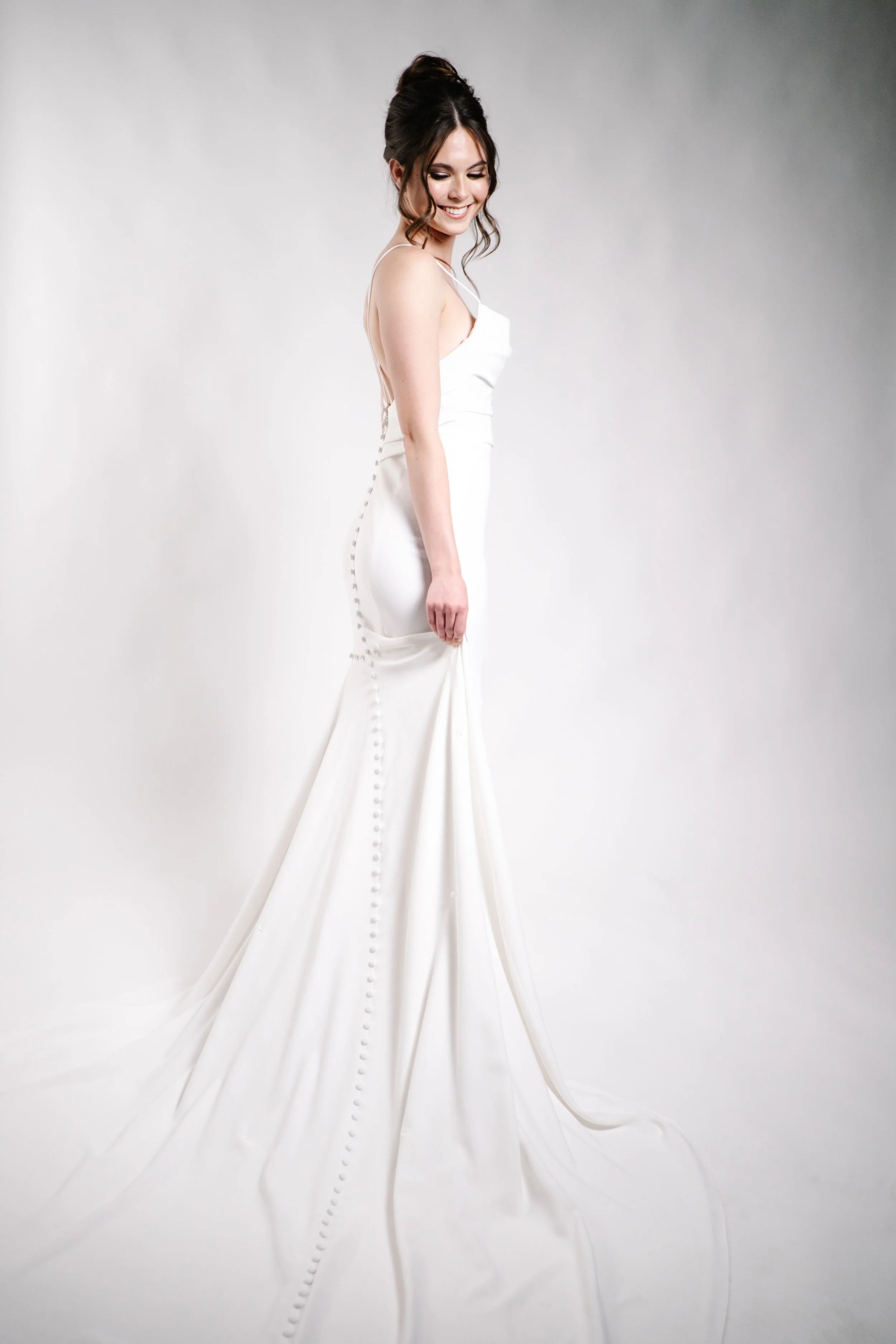 A woman wearing a white wedding dress with a train, standing against a plain white background, smiling and looking down.