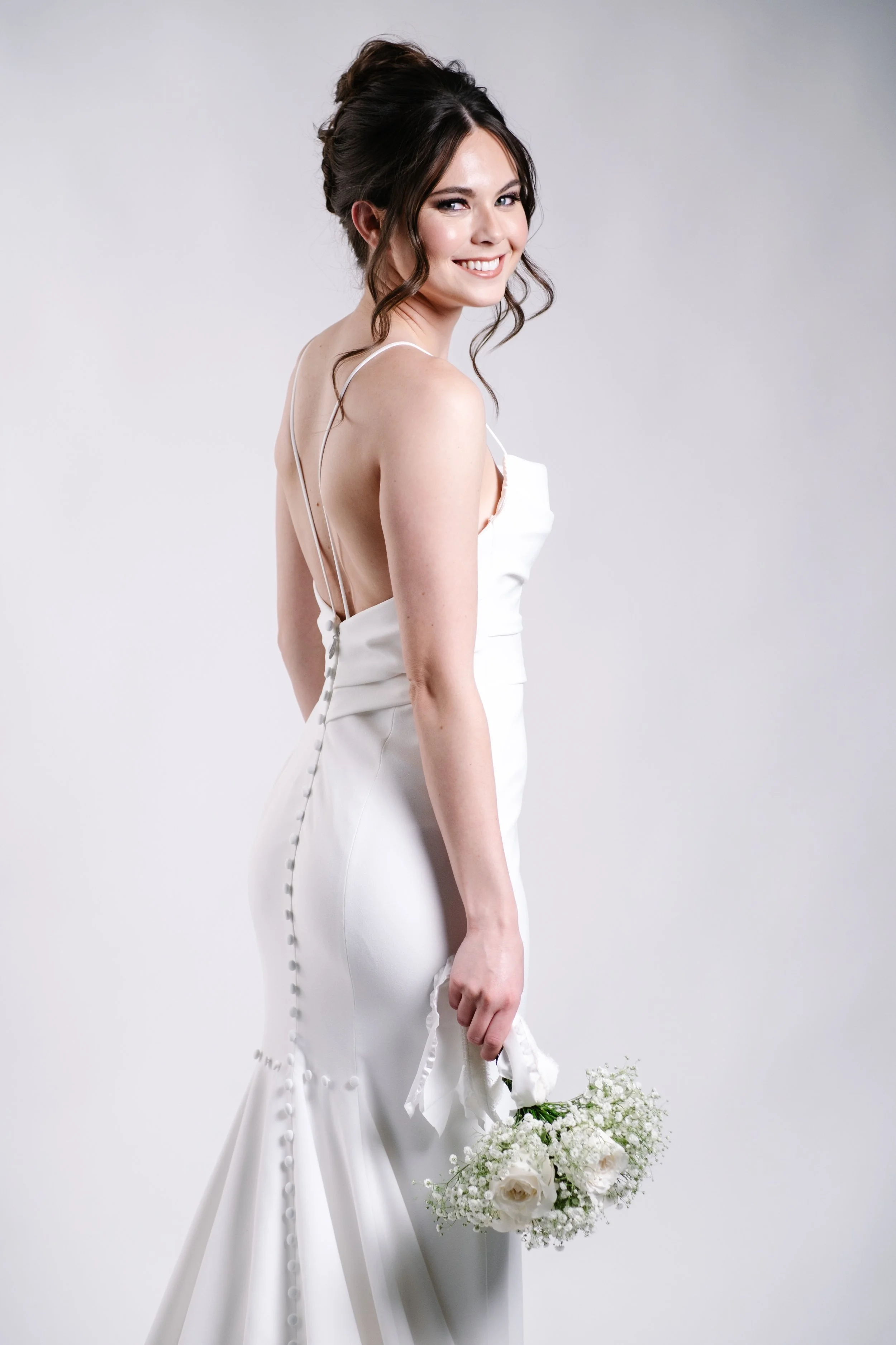 A smiling woman in a white wedding dress holding a bouquet of white roses and baby's breath, standing against a plain white background.