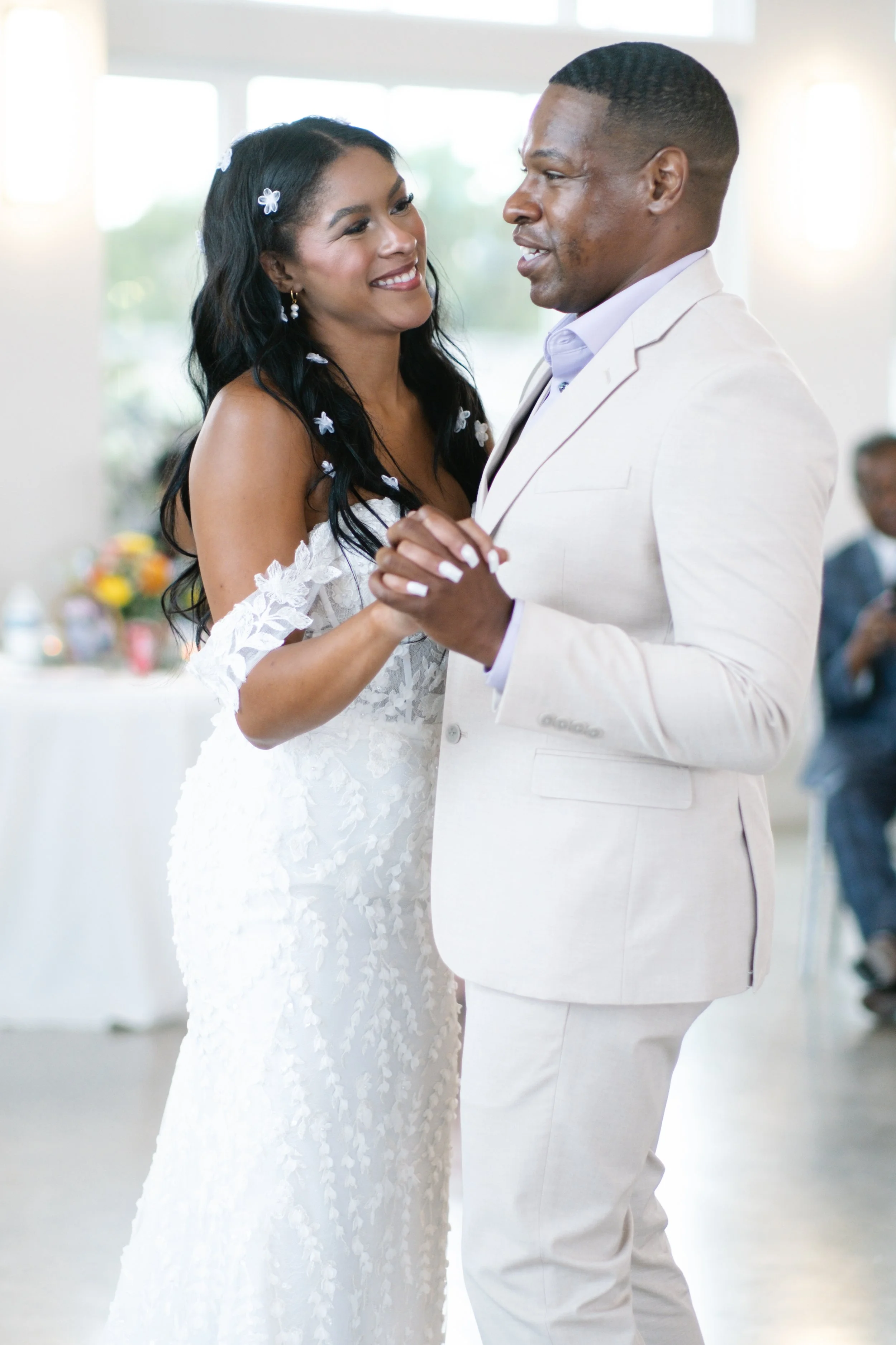 A couple dancing at their wedding reception, the woman in a white wedding dress with floral details and the man in a light-colored suit.